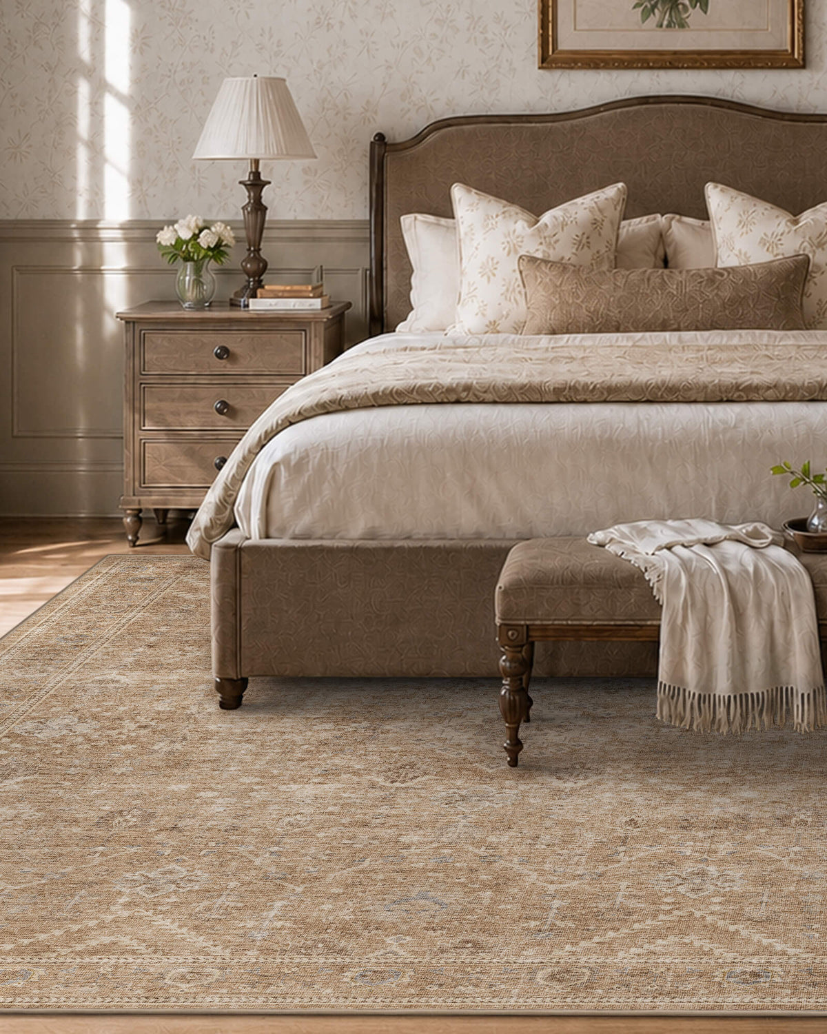 A beige vintage washable area rug with a softly distressed floral pattern and delicate ornamental border adds timeless elegance to a traditional bedroom with an upholstered bed, layered cream bedding, antique wood furniture, and warm natural light.