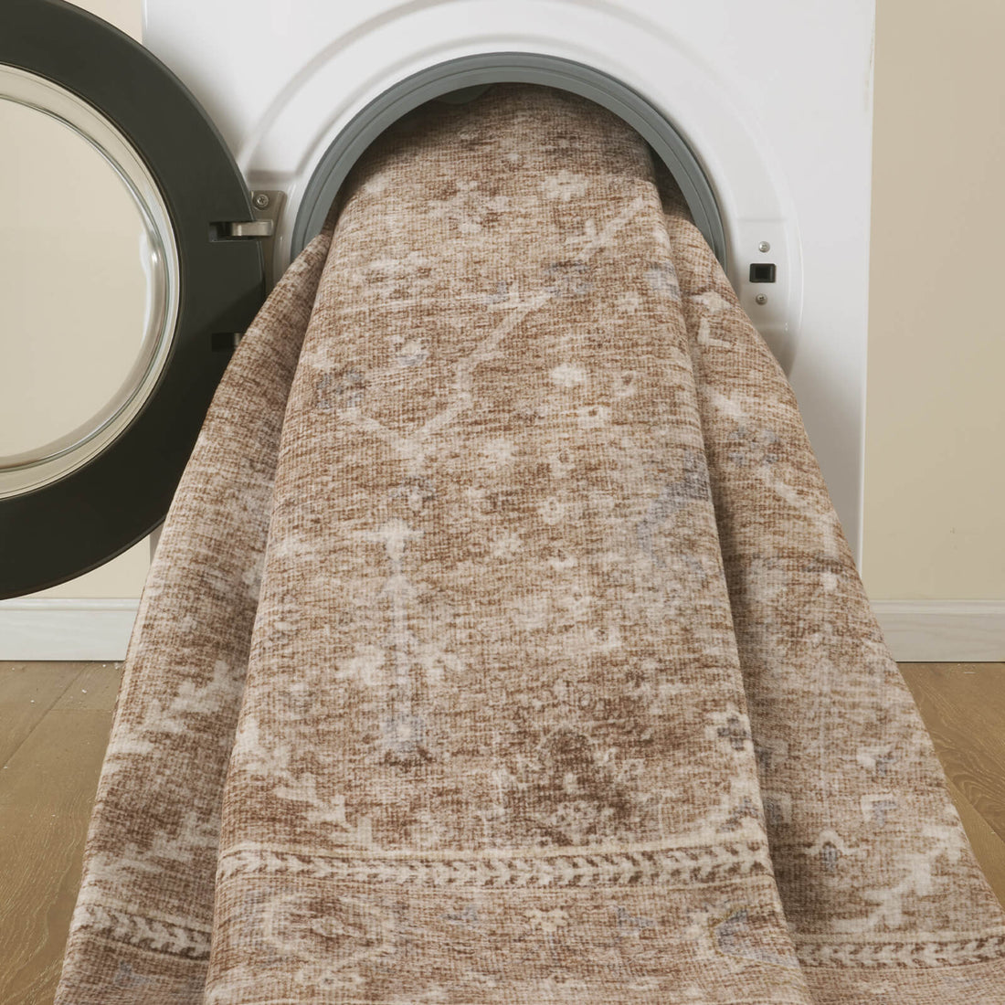 A beige vintage washable area rug is shown being placed in a front-loading washing machine, highlighting its easy-care design, softly distressed floral pattern, and warm neutral tones for busy family homes, living rooms, and bedrooms.