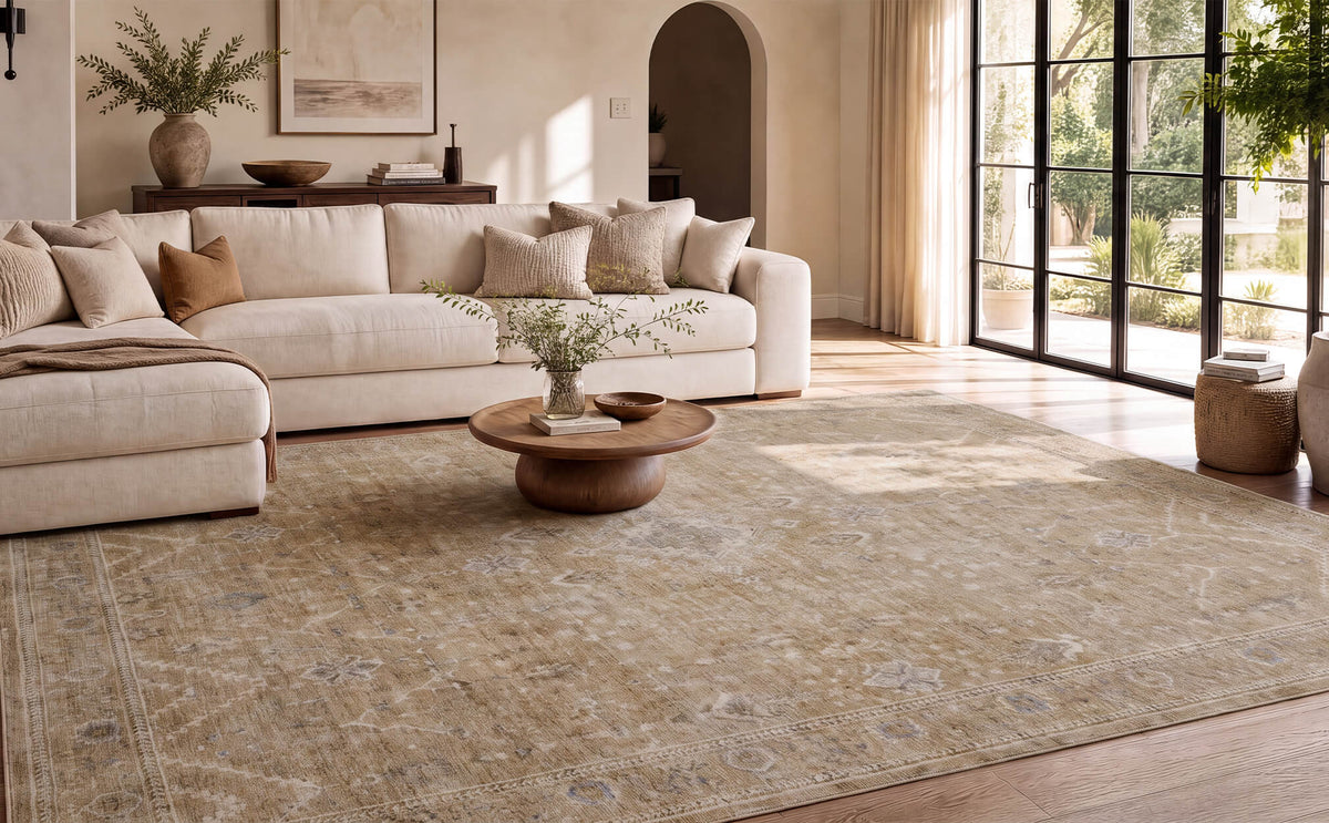 A beige vintage washable area rug with a softly distressed floral pattern and subtle ornamental details adds timeless elegance to a spacious living room with a cream sectional sofa, round wood coffee table, floor-to-ceiling windows, and warm natural sunlight.