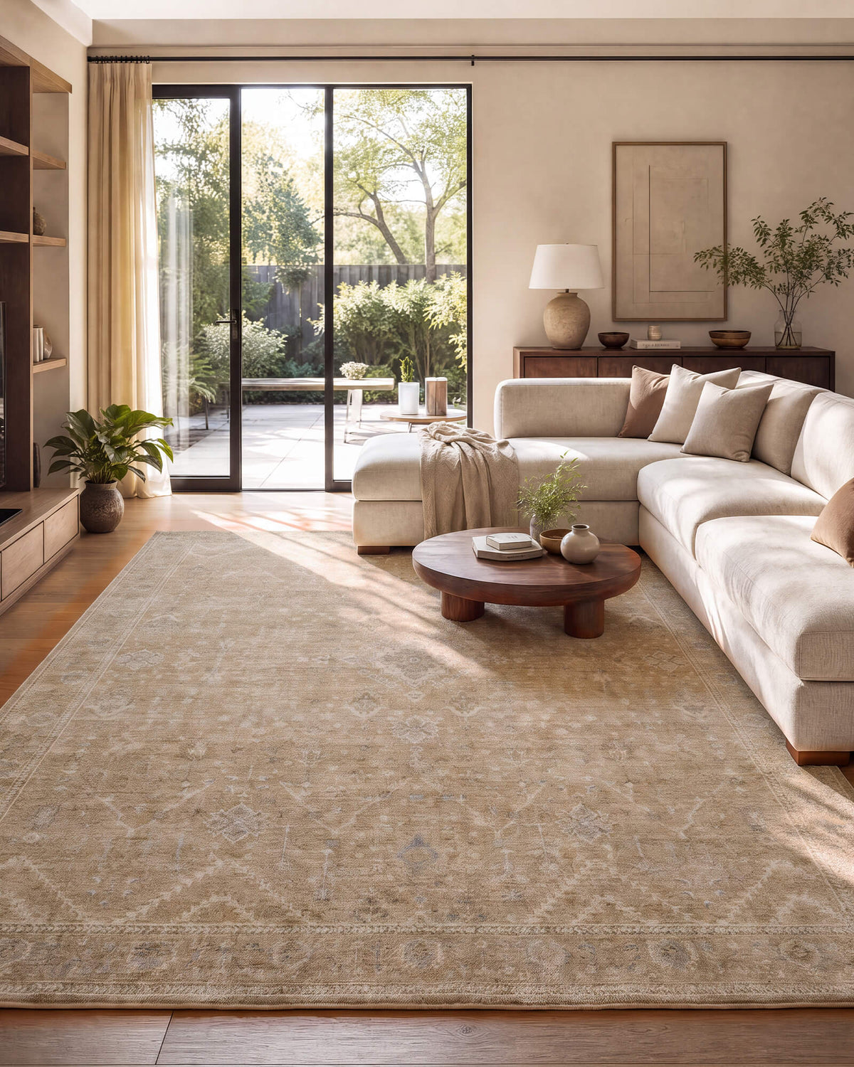 A beige vintage washable area rug with a softly distressed floral pattern and delicate ornamental border brings understated elegance to a sunlit modern living room with a cream sectional sofa, round wood coffee table, floor-to-ceiling windows, and warm neutral decor.