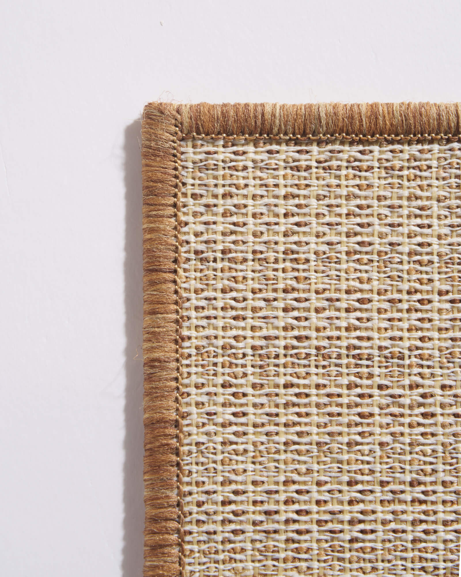 A close-up view of a brown abstract outdoor rug showing its woven grid backing, reinforced corner edge, and durable construction for weather-resistant use on patios, decks, porches, and outdoor living areas.