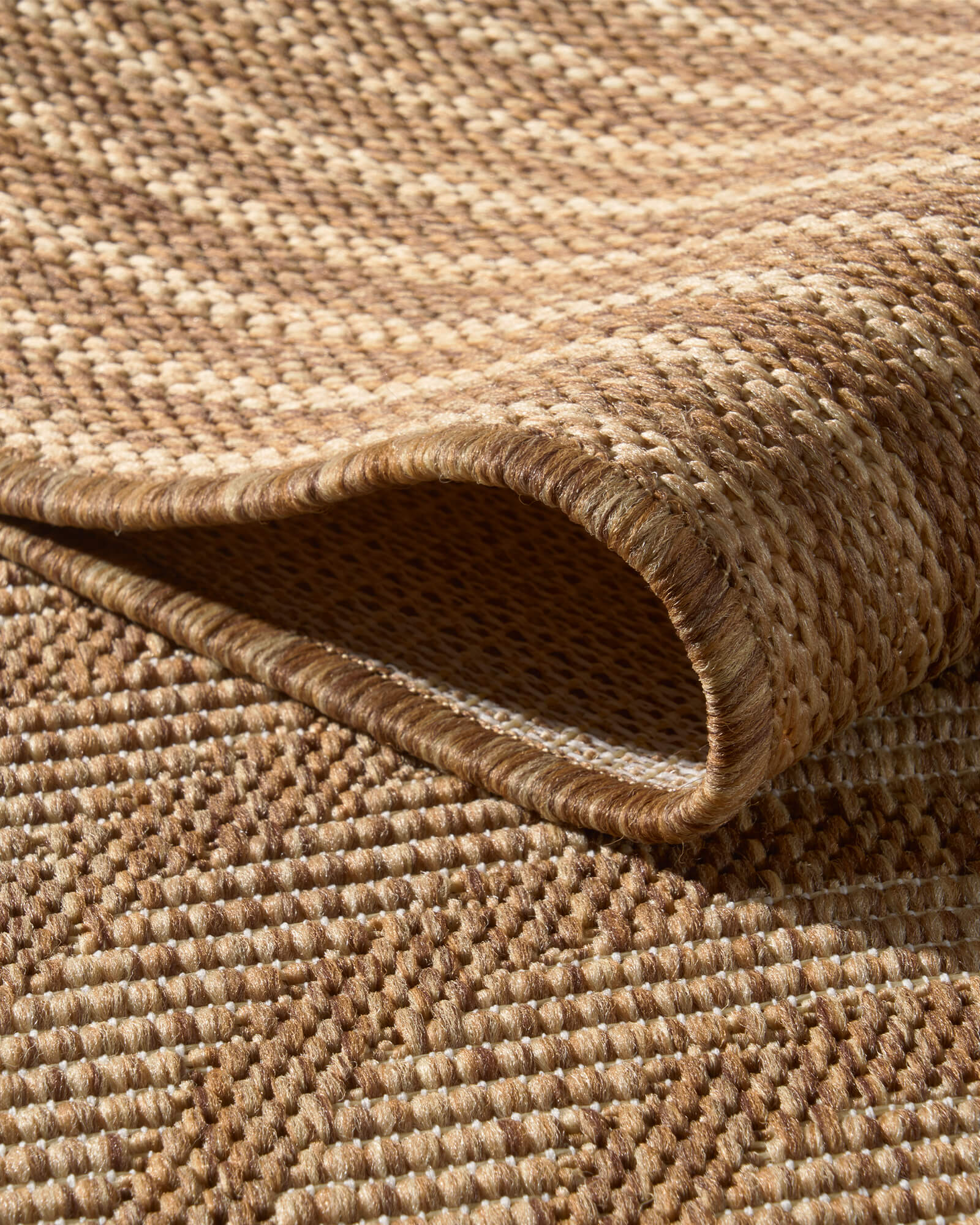 A close-up view of a brown abstract outdoor rug showing its curved fold, woven texture, bound edge, and raised arched geometric pattern for durable patio, deck, and porch styling.