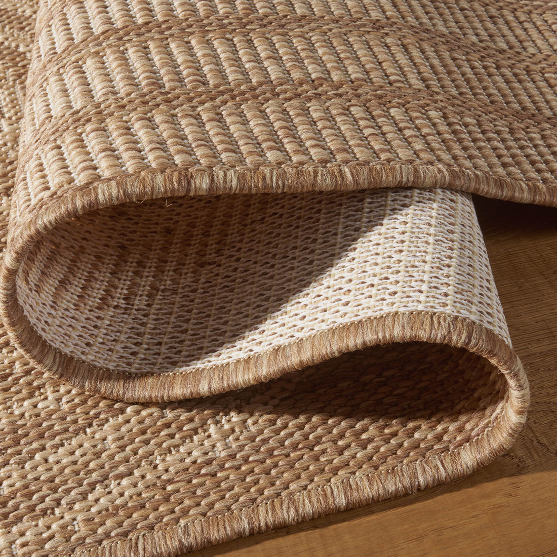 A close-up view of a brown abstract outdoor rug showing its folded corner, woven backing, bound edge, and raised arch line texture for durable weather-resistant use on patios, decks, and porches.