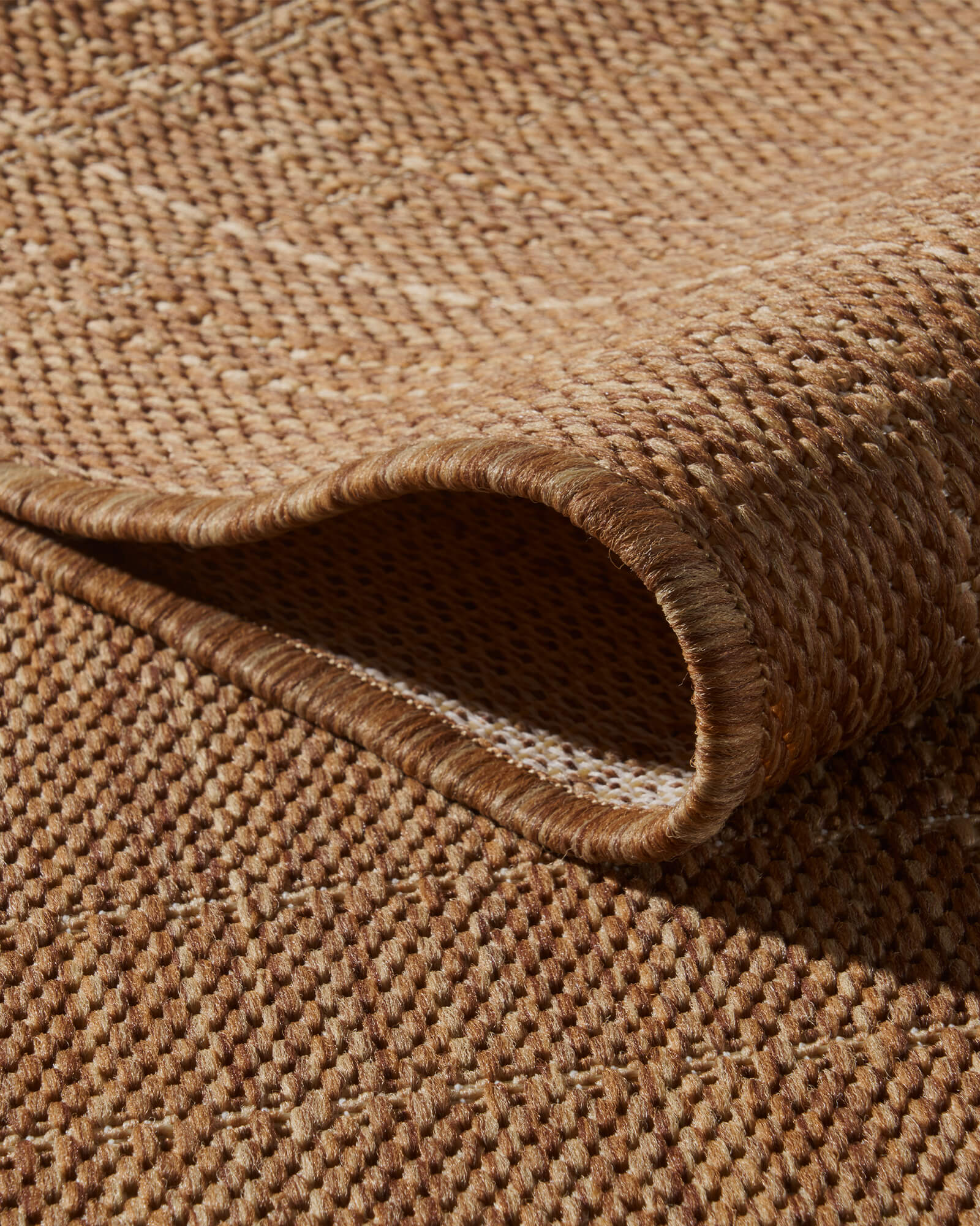 A close-up view of a brown plain weave outdoor rug showing its folded corner, open backing, raised grid texture, and durable bound edge for patios, decks, porches, and outdoor living spaces.