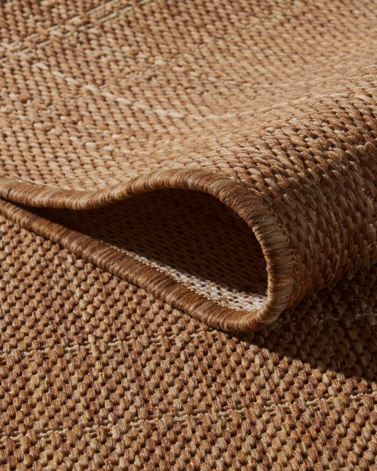 A close-up view of a brown plain weave outdoor rug showing its folded corner, open backing, raised grid texture, and durable bound edge for patios, decks, porches, and outdoor living spaces.