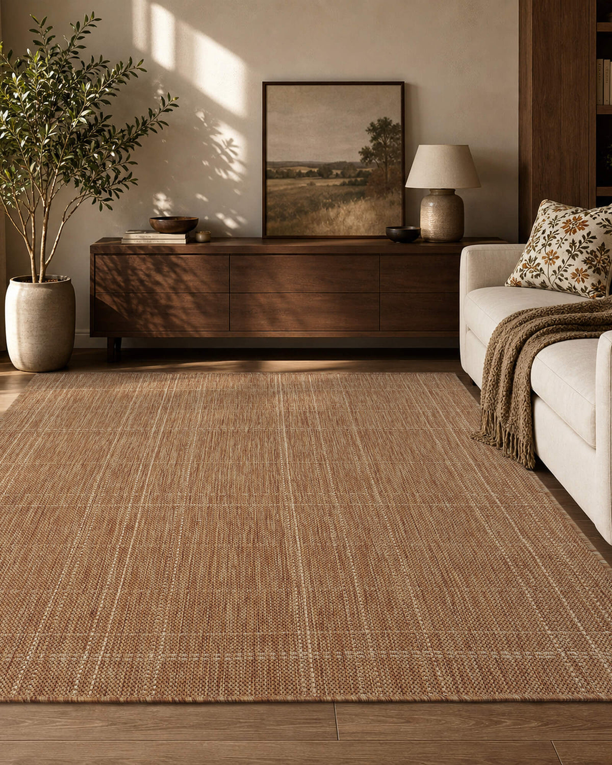 A brown plain weave indoor outdoor area rug with a subtle check border adds warm natural texture to a cozy living room with a wood console, cream sofa, soft sunlight, and earthy home decor.