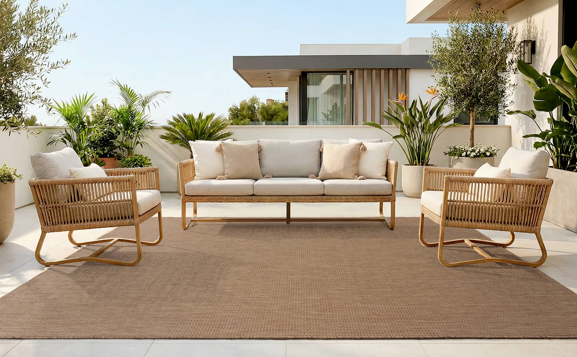 A brown plain weave outdoor area rug adds warm minimal texture to a modern patio seating area with woven chairs, a neutral sofa, tropical greenery, and weather-resistant outdoor style.