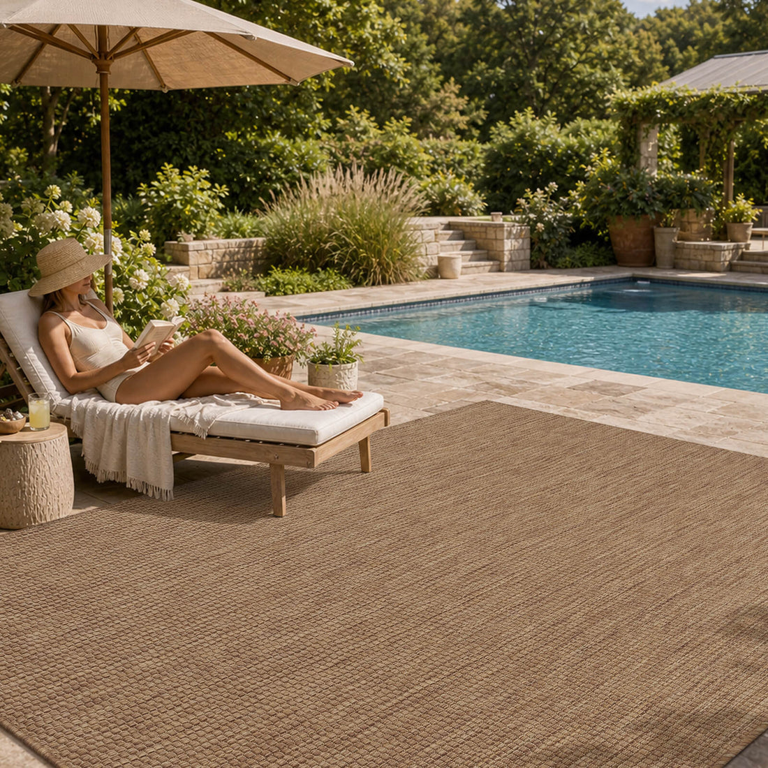 A brown plain weave outdoor area rug adds natural woven texture to a poolside patio with a chaise lounge, umbrella shade, garden greenery, and relaxed summer outdoor decor.