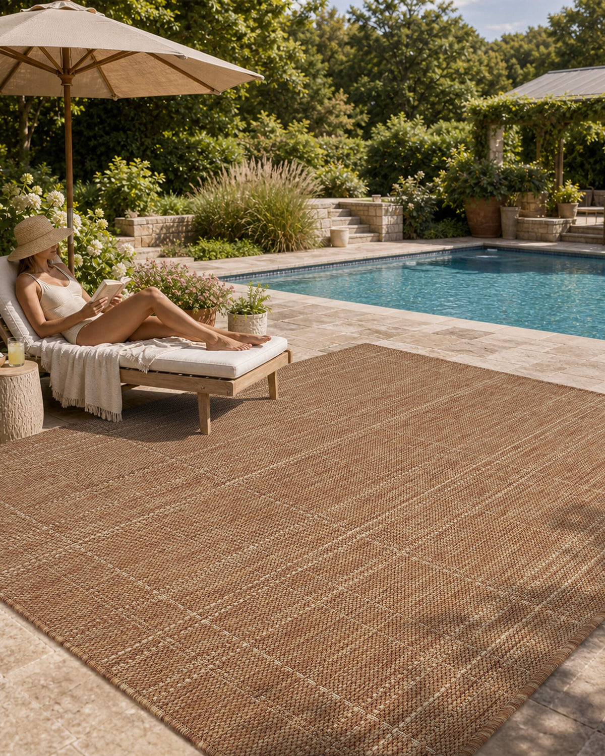 A brown plain weave outdoor area rug with a subtle check border adds natural woven texture to a poolside patio with a chaise lounge, umbrella shade, garden greenery, and relaxed summer style.
