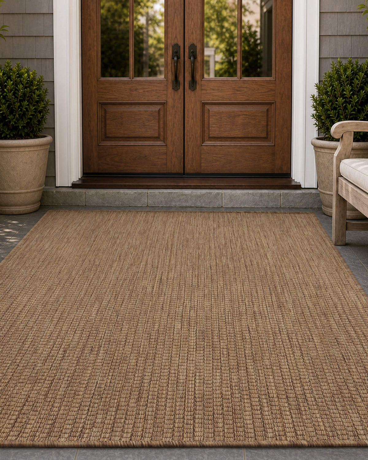 A brown plain weave outdoor rug adds natural texture to a front door entryway, creating a warm weather-resistant porch look with wood double doors, potted greenery, and classic outdoor styling.