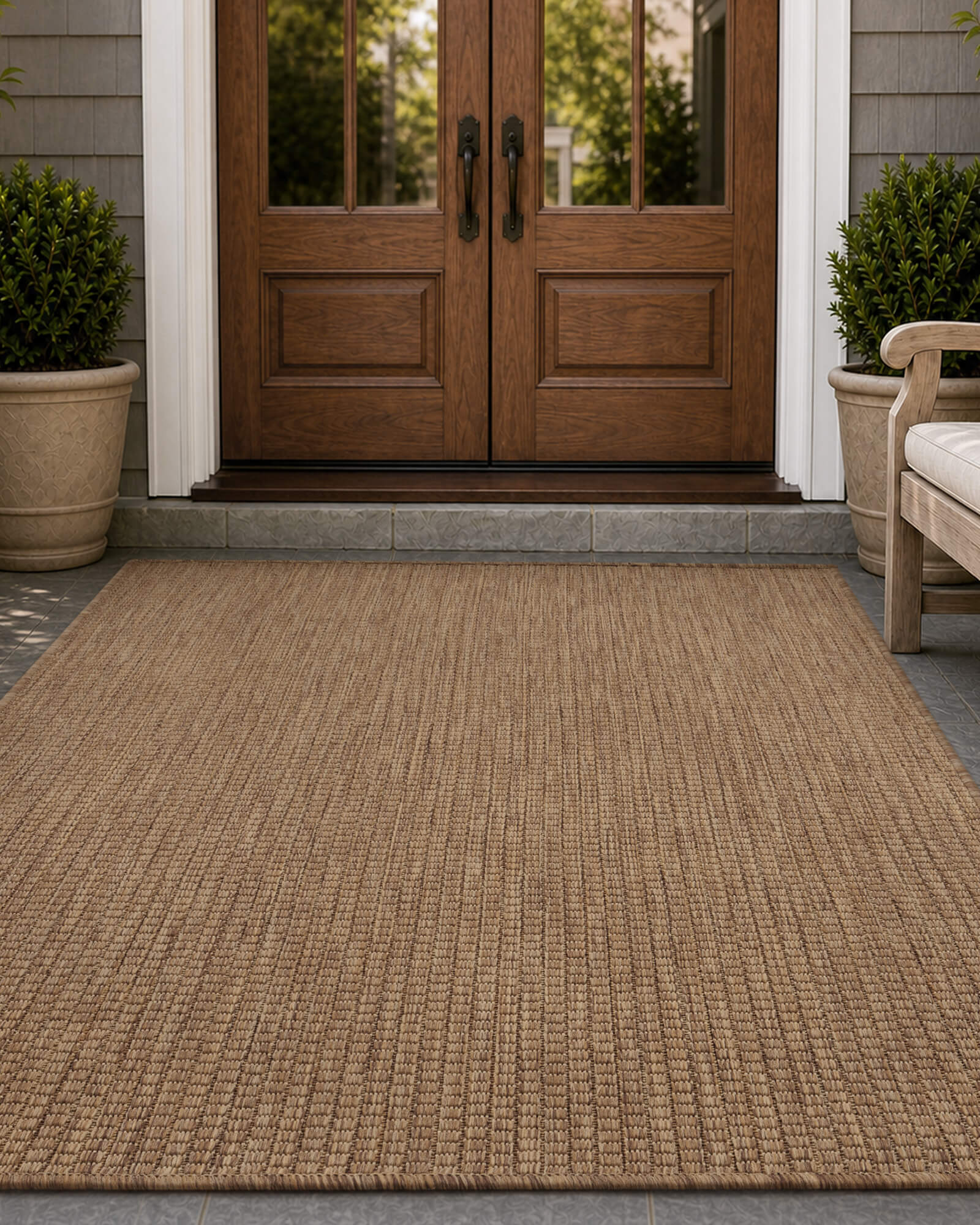 A brown plain weave outdoor rug adds natural texture to a front door entryway, creating a warm weather-resistant porch look with wood double doors, potted greenery, and classic outdoor styling.