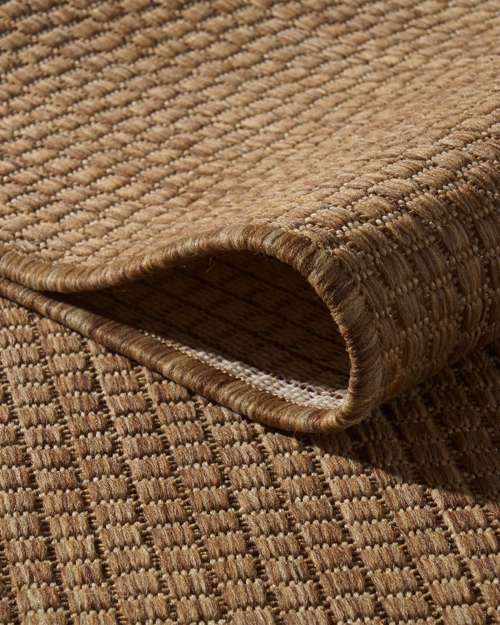 A close-up view of a brown plain weave outdoor rug showing its looped corner, basketweave surface, bound edge, and durable woven construction for patios, decks, porches, and outdoor living spaces.