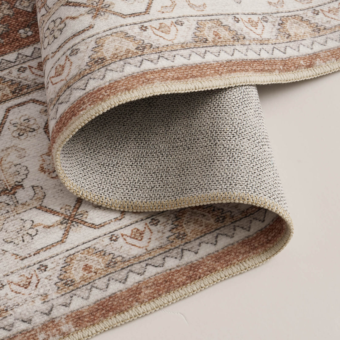 A close-up view of a brown vintage washable area rug showing its folded corner, gray non-slip backing, distressed ornate border, and neatly finished edge for cozy living room and bedroom use.