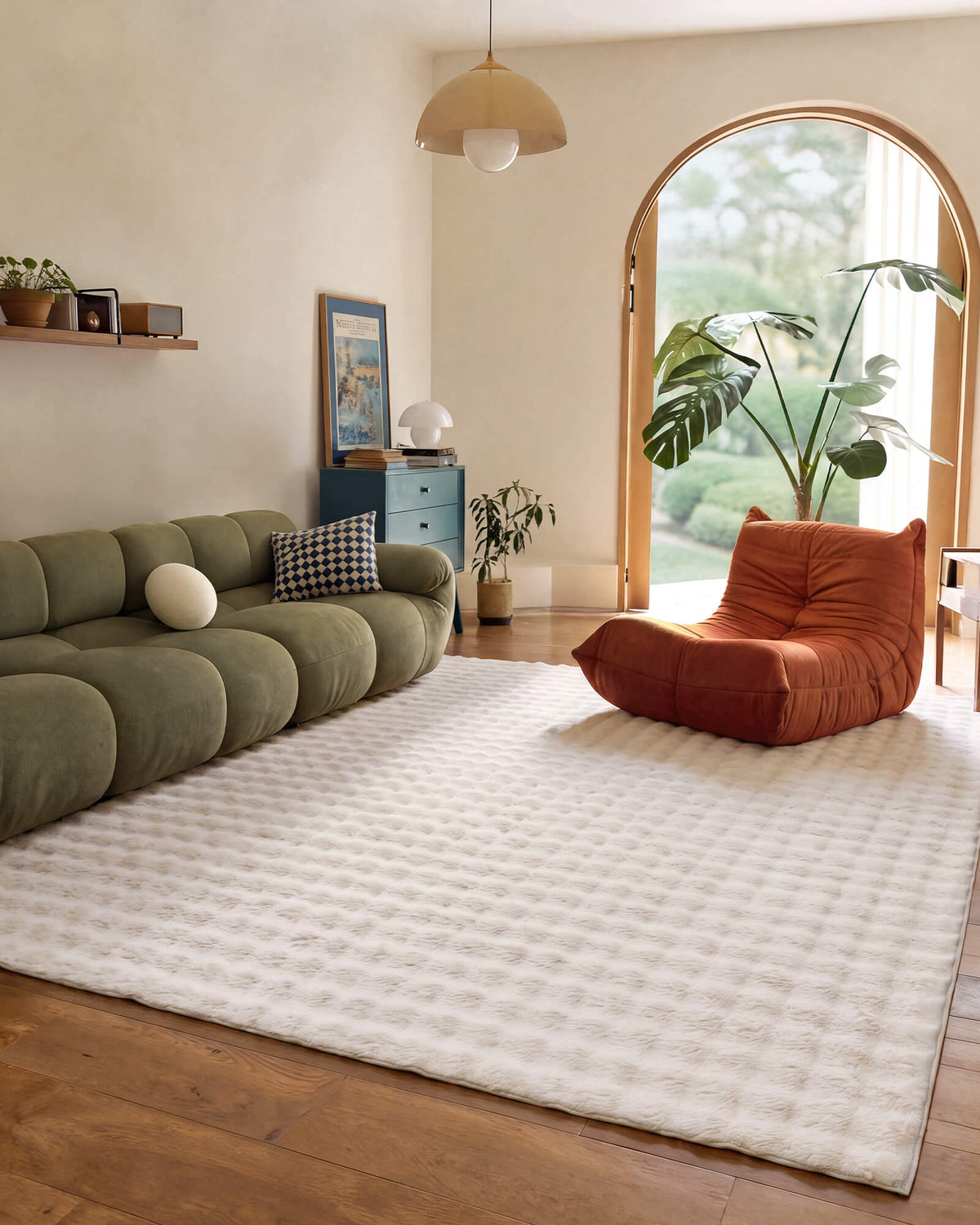 A cream Cloud Soft shag area rug with an ultra-fluffy long-pile surface and plush checkered texture brings cloud-like softness and luxurious comfort to a modern living room with a sculptural olive green sofa, rust orange lounge chair, arched window, and warm natural sunlight.