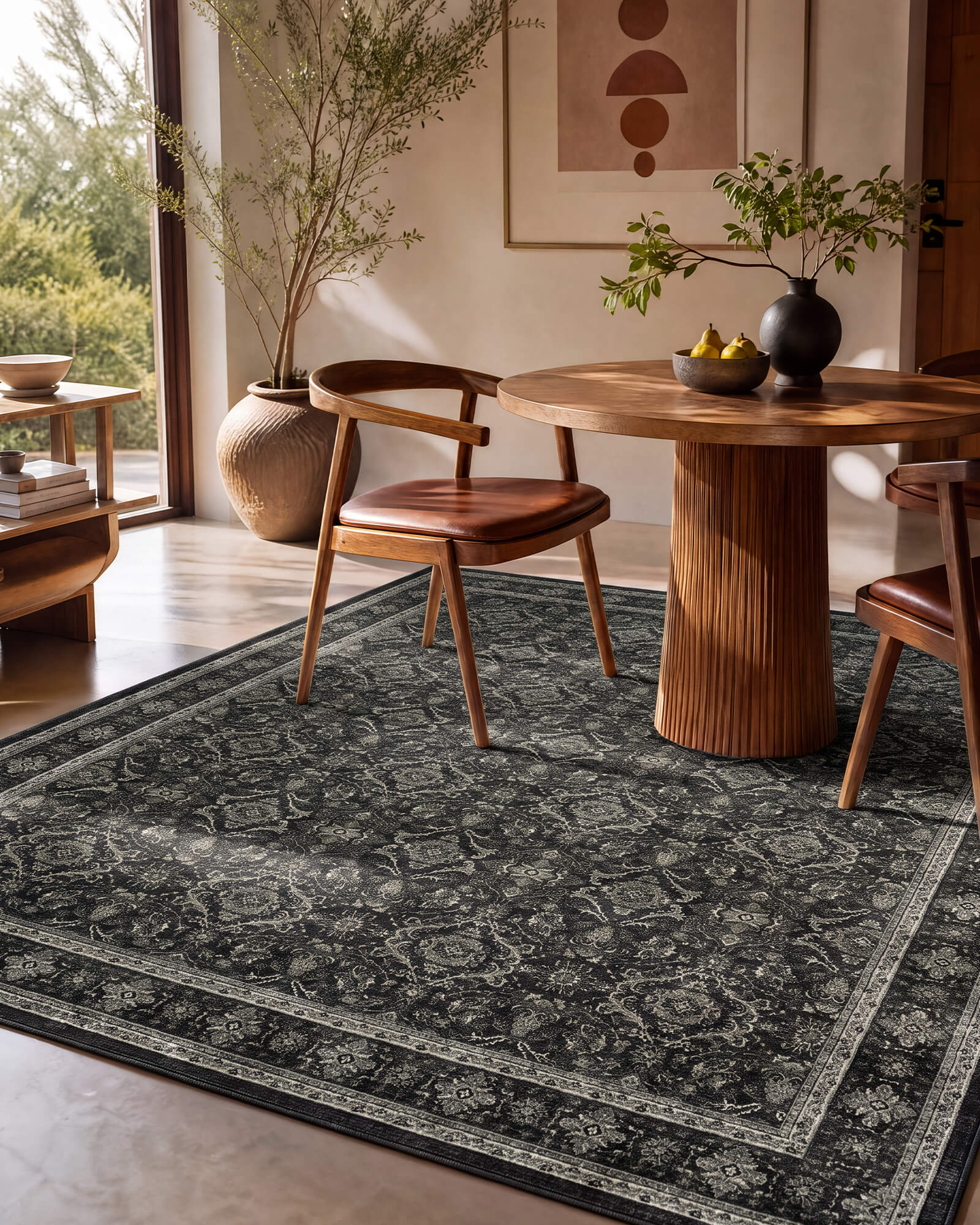 A dark charcoal vintage washable rug with a faded ornamental border pattern adds rich contrast to a warm dining room with a round wood table, leather chairs, natural textures, and sunlit modern decor.