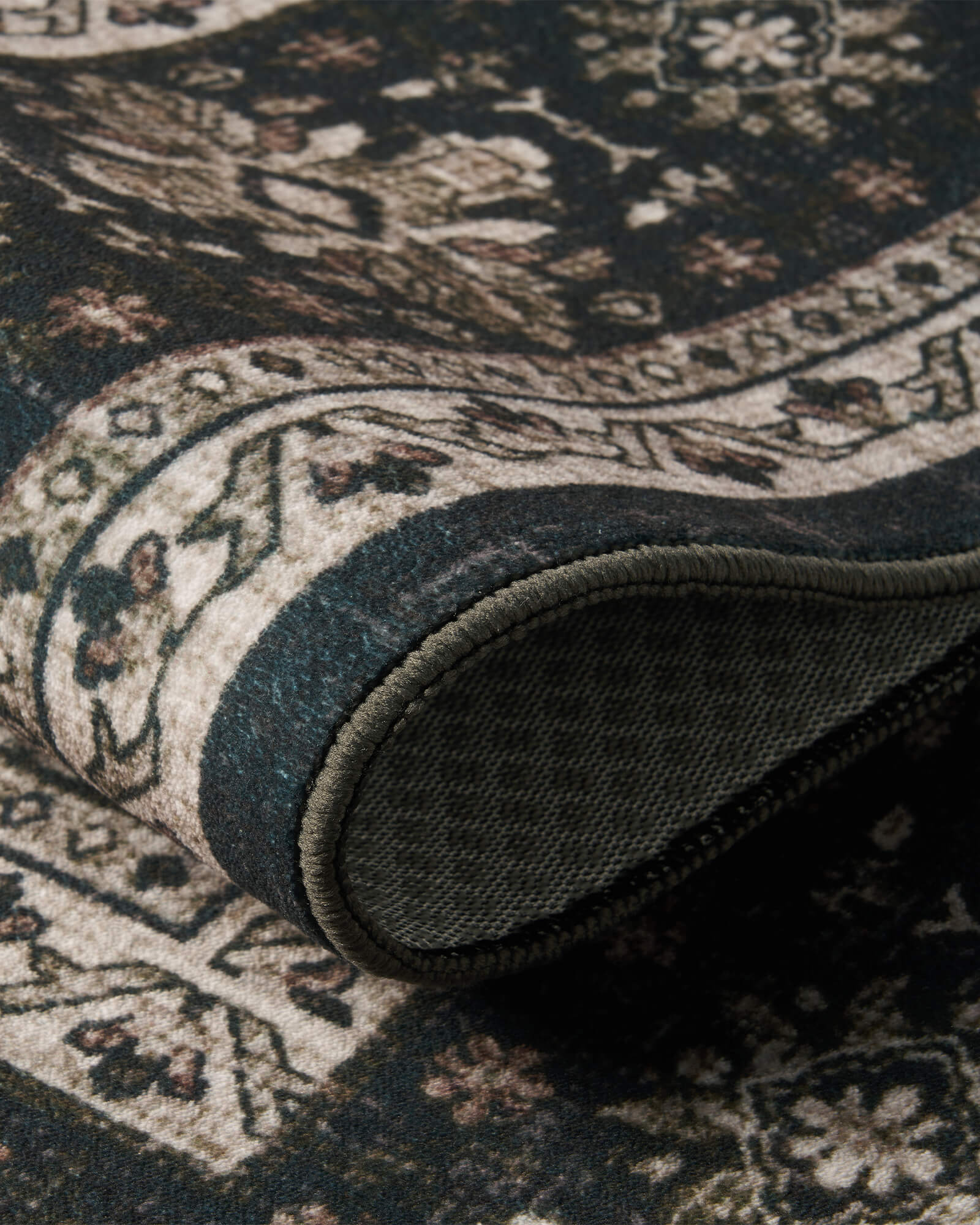A close-up view of a dark charcoal vintage washable rug showing its curled corner, non-slip backing, faded floral border, and soft low-pile texture for easy everyday use in living rooms, bedrooms, and cozy home spaces.