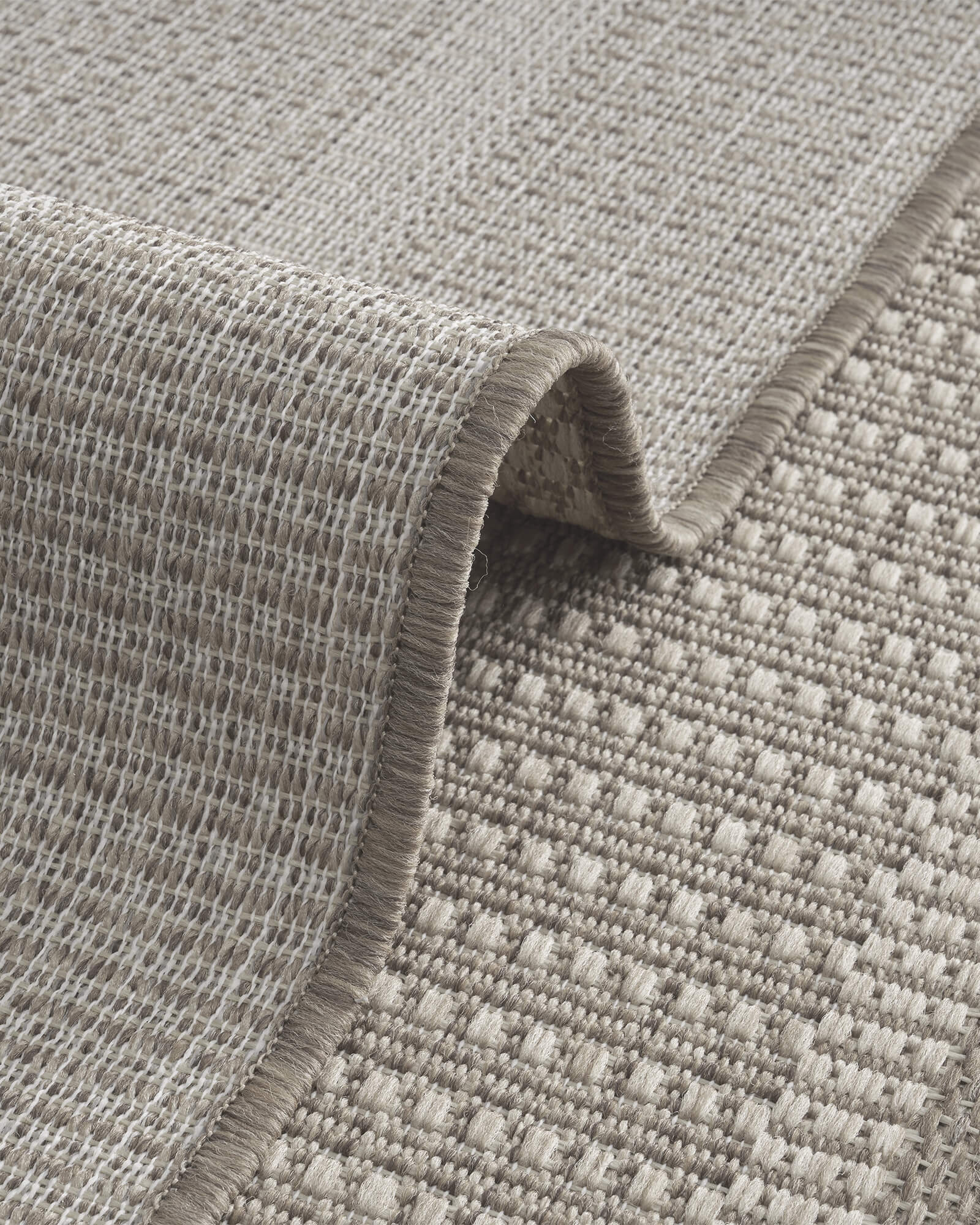 A foldable gray woven indoor outdoor rug features a durable jute-look texture and reinforced edge for easy-care use on patios, porches, kitchens, and indoor living spaces.