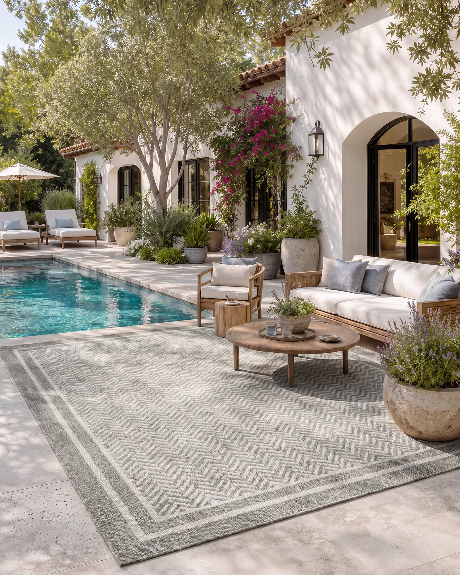 A gray chevron indoor outdoor rug adds durable woven texture and easy-care style to a poolside patio lounge with outdoor seating, garden greenery, and warm Mediterranean-inspired decor.