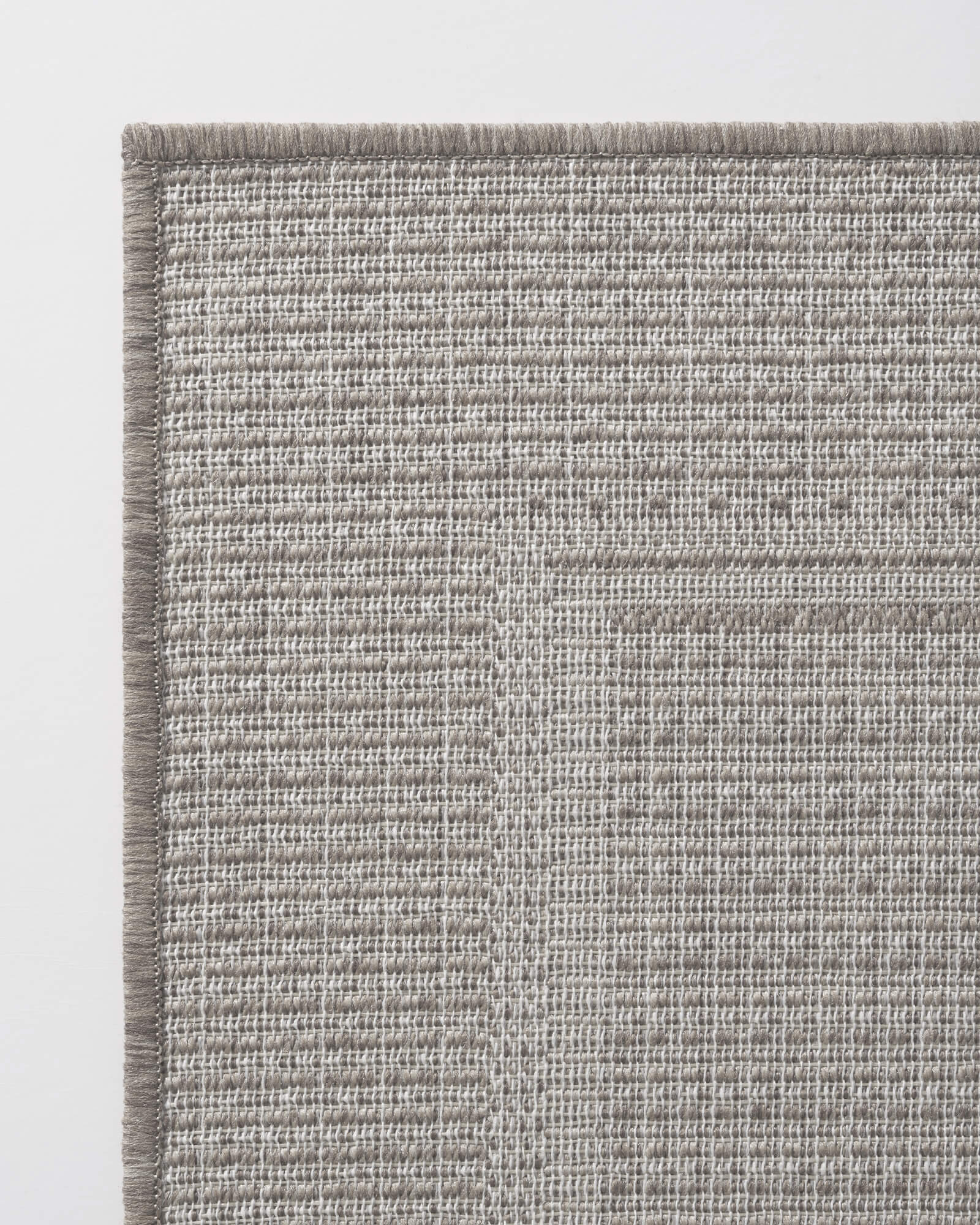 A gray woven indoor outdoor rug features a durable jute-look texture and reinforced border edge, bringing easy-care style to patios, porches, sunrooms, kitchens, and high-traffic spaces.
