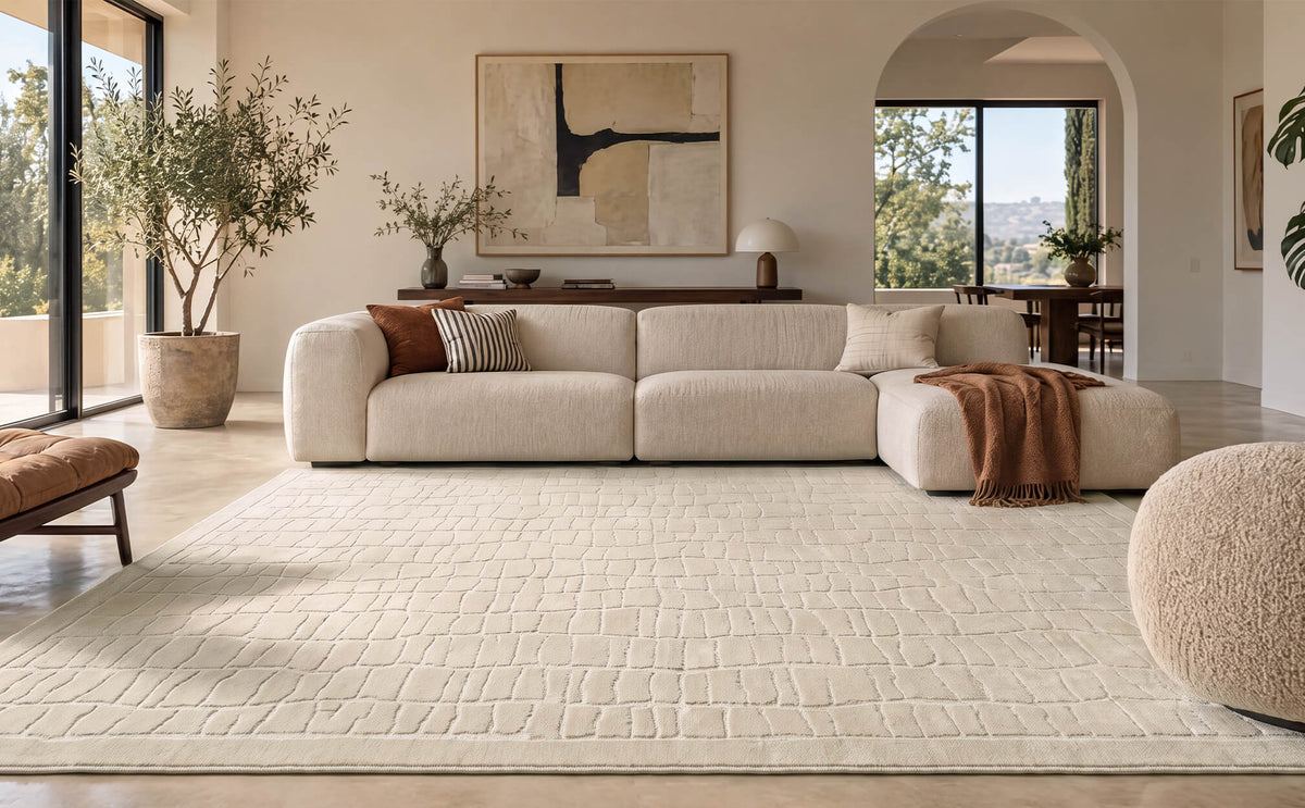 An ivory luxury Wilton jacquard area rug with a raised block texture adds sculptural softness and refined dimension to a wide modern living room with a cream sectional sofa, arched doorway, curated artwork, and warm natural light.