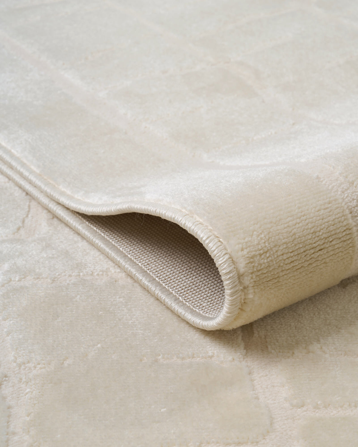 A close-up view of an ivory luxury Wilton jacquard rug showing its curled edge, plush raised block pattern, structured backing, and refined bound finish for premium living room and bedroom decor.