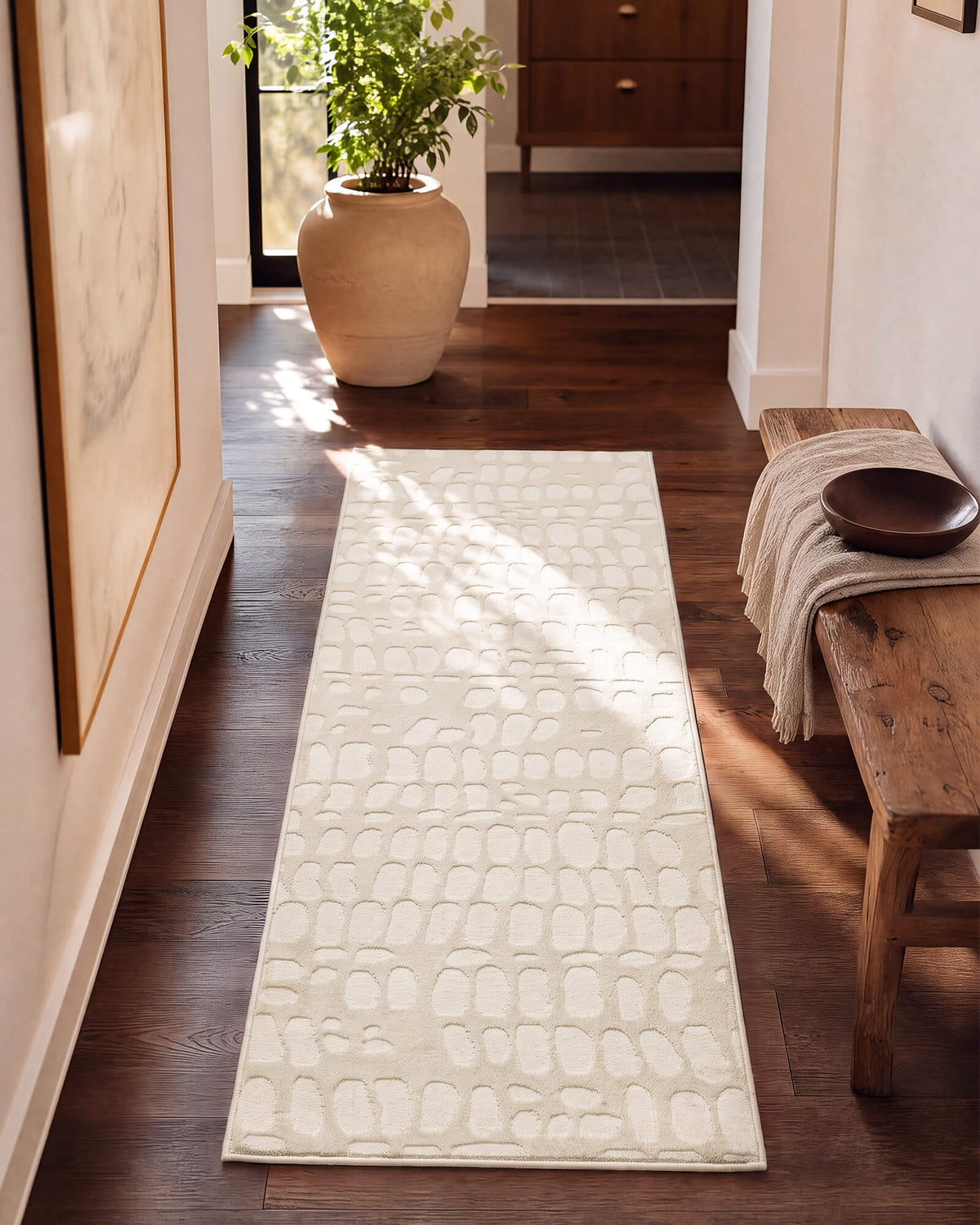 An ivory luxury Wilton jacquard runner rug with raised pebble-inspired texture adds sculptural softness to a sunlit entryway hall with warm wood floors, a rustic bench, potted greenery, and refined neutral decor.