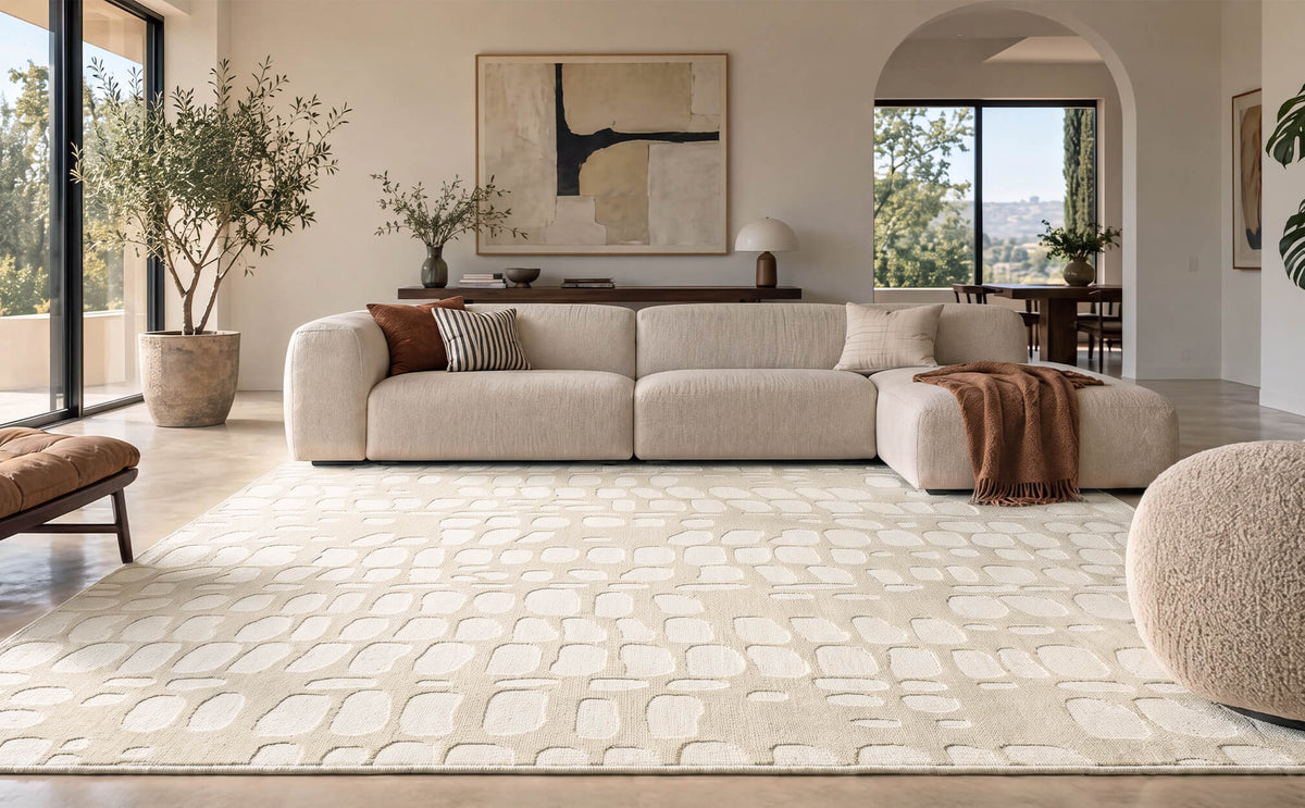 An ivory premium Wilton jacquard area rug with a raised pebble-inspired pattern brings sculptural texture and refined softness to a luxury living room with a curved sectional sofa, arched doorway, curated artwork, and warm modern neutral decor.