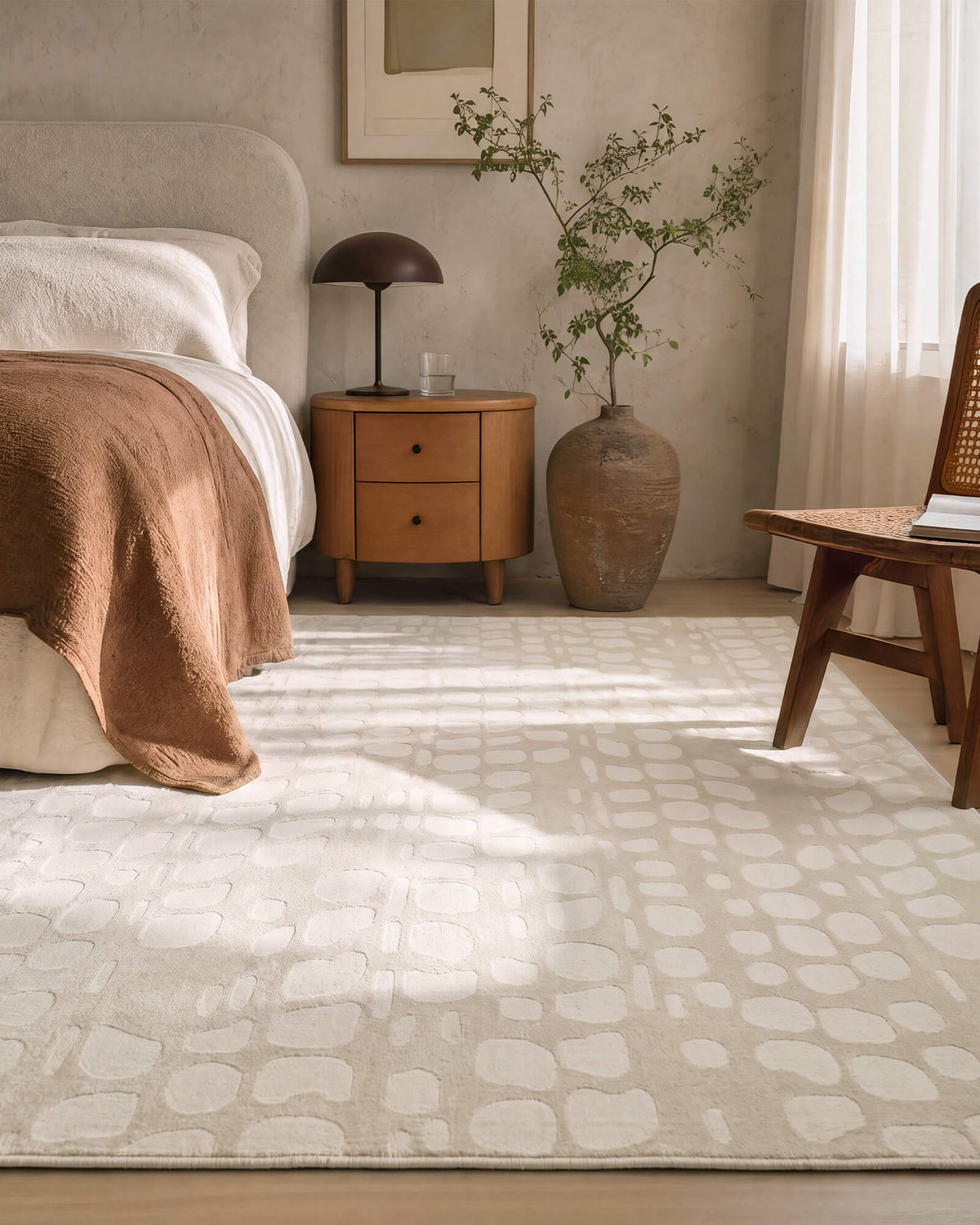 An ivory premium Wilton jacquard area rug with a raised pebble-inspired pattern adds sculptural texture and refined softness to an elegant bedroom with warm wood furniture, layered bedding, and natural sunlight.