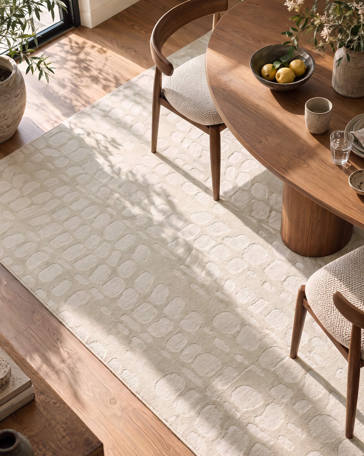 An ivory premium Wilton jacquard rug with raised pebble-inspired texture adds sculptural softness to a refined dining room with a round wood table, natural sunlight, and elevated warm wood decor.
