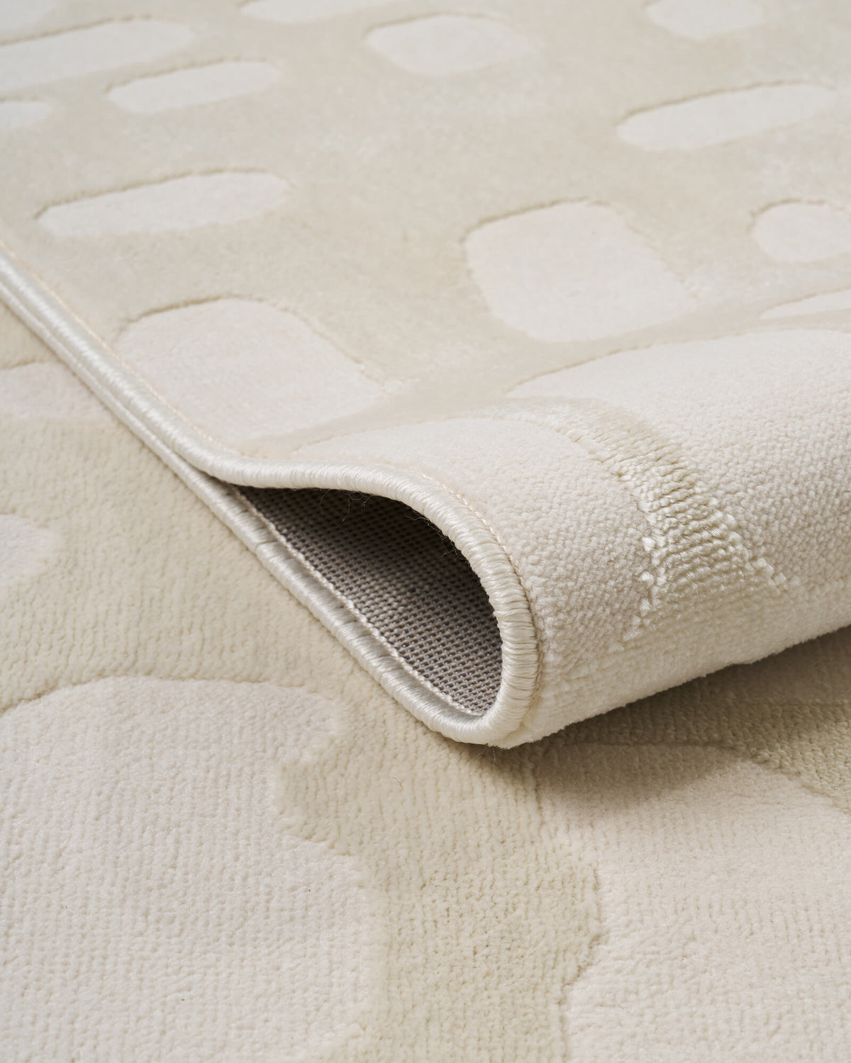 A close-up view of an ivory premium Wilton jacquard rug showing its curled edge, woven backing, raised pebble-inspired texture, and refined bound finish for elevated living room and bedroom decor.