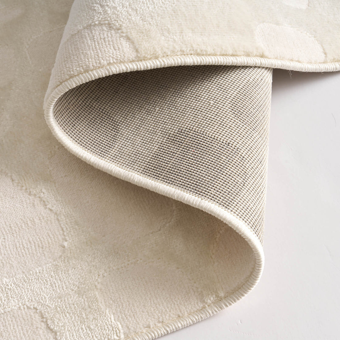 A close-up view of an ivory premium Wilton jacquard rug showing its folded corner, woven backing, raised pebble-inspired texture, and refined bound edge for elevated living room and bedroom decor.