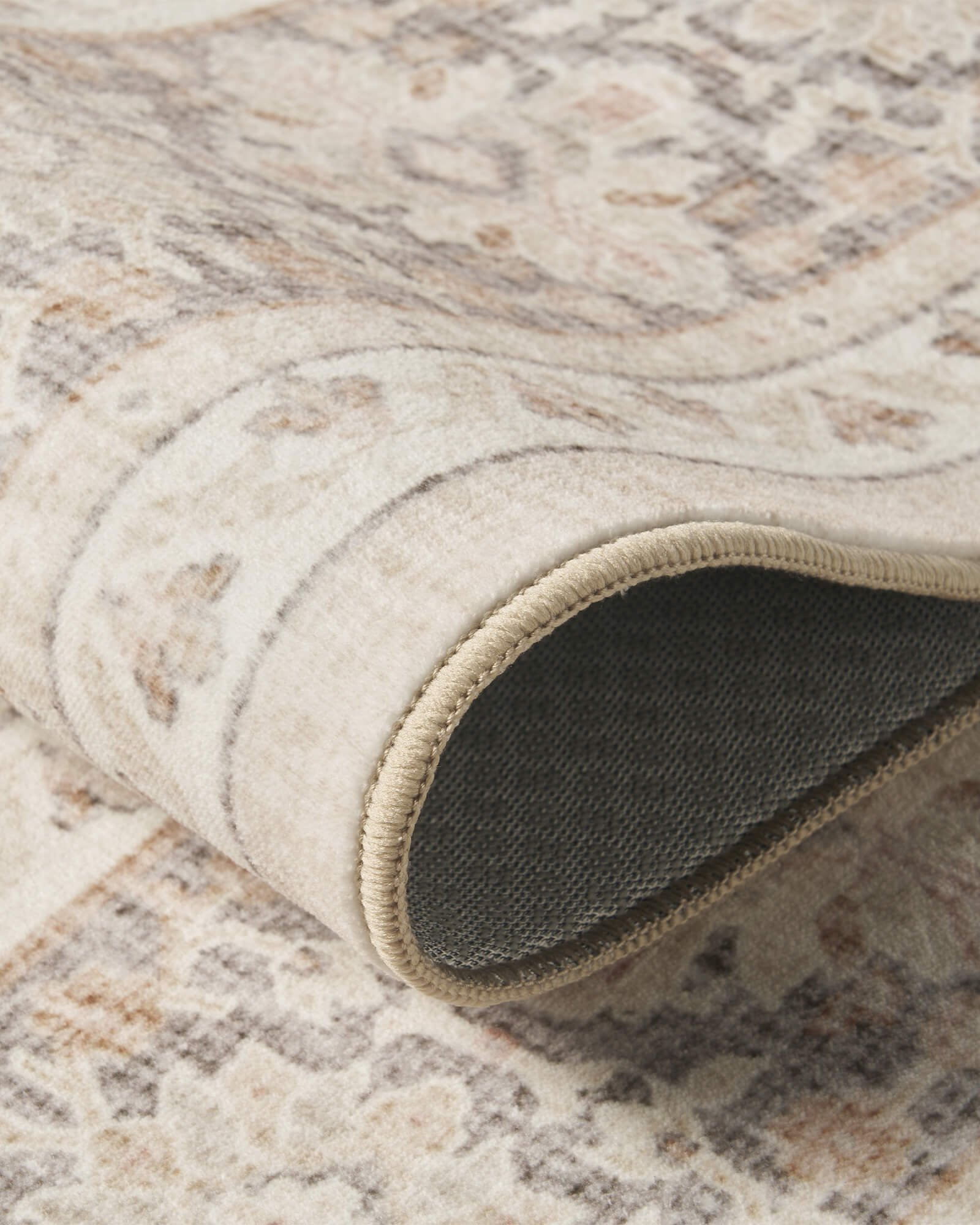 A close-up view of an ivory vintage washable rug showing its rolled edge, gray backing, faded medallion pattern, and flexible low-pile construction for easy everyday use in living rooms and bedrooms.