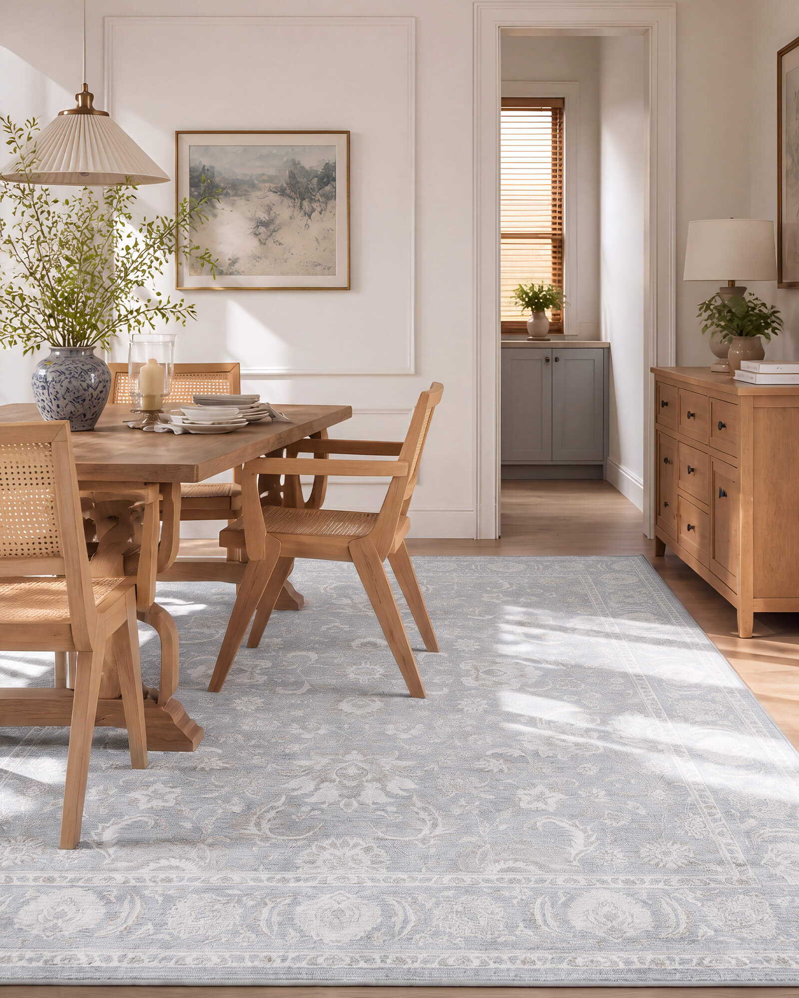 A light blue vintage washable rug with a soft ornamental border pattern adds airy elegance to a sunlit dining room with a wood table, cane chairs, warm natural light, and classic cottage-inspired decor.