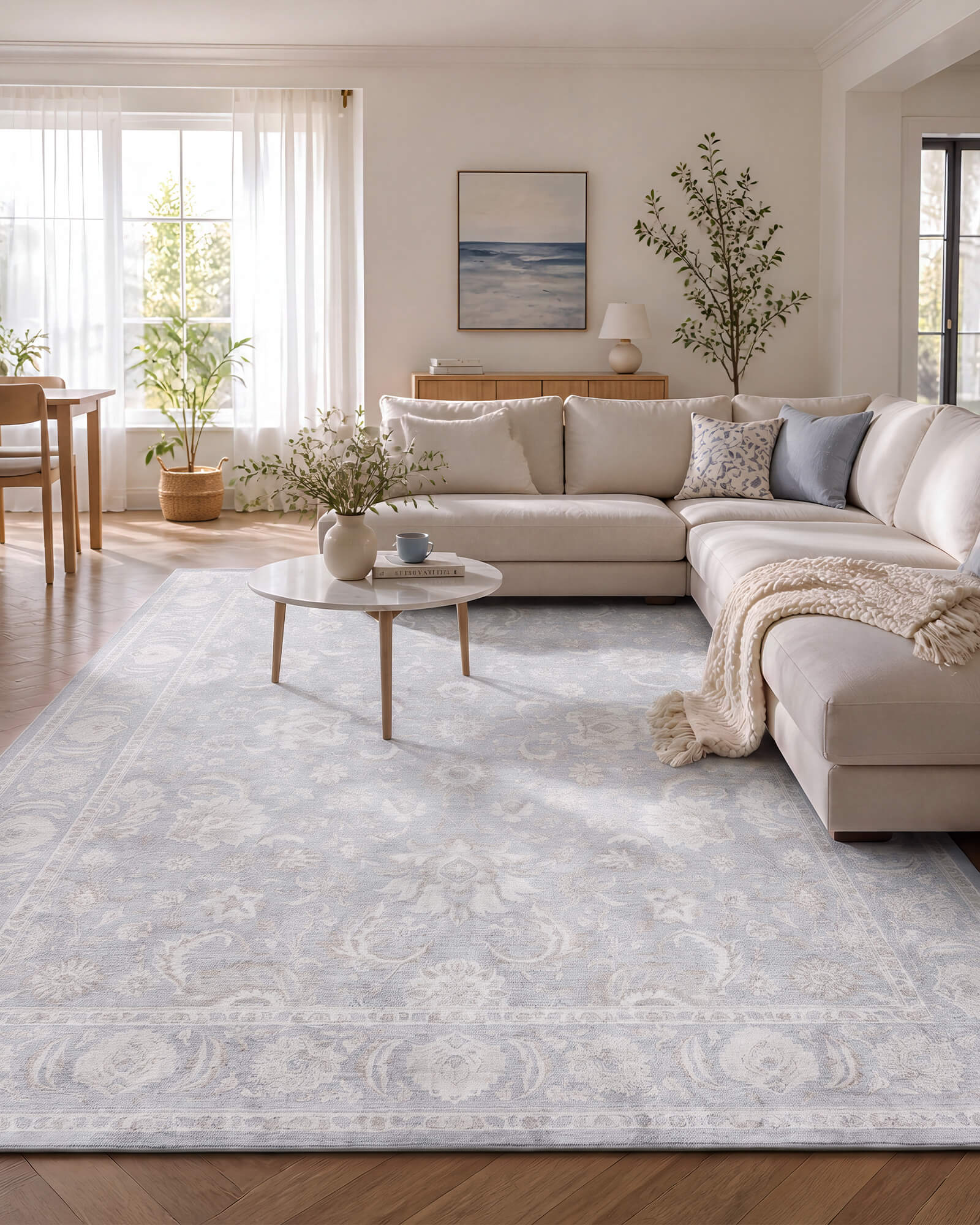 A light blue vintage washable rug with a soft ornamental border pattern brightens an airy living room with a sectional sofa, natural wood floors, sheer curtains, and cozy neutral decor.
