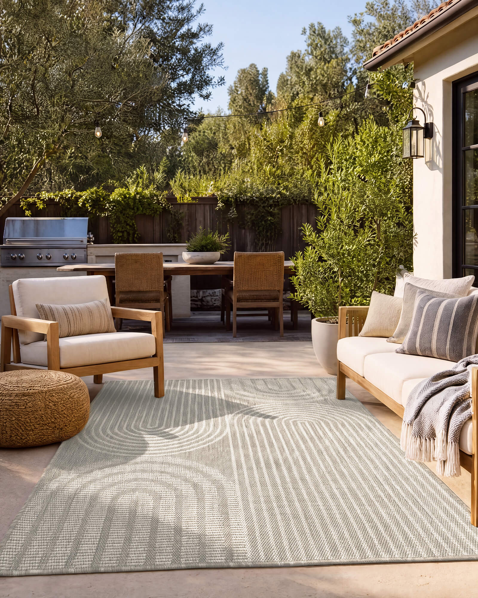 A light gray abstract indoor outdoor rug with a modern arch line pattern adds soft texture to a backyard patio lounge with outdoor seating, greenery, string lights, and weather-resistant style.