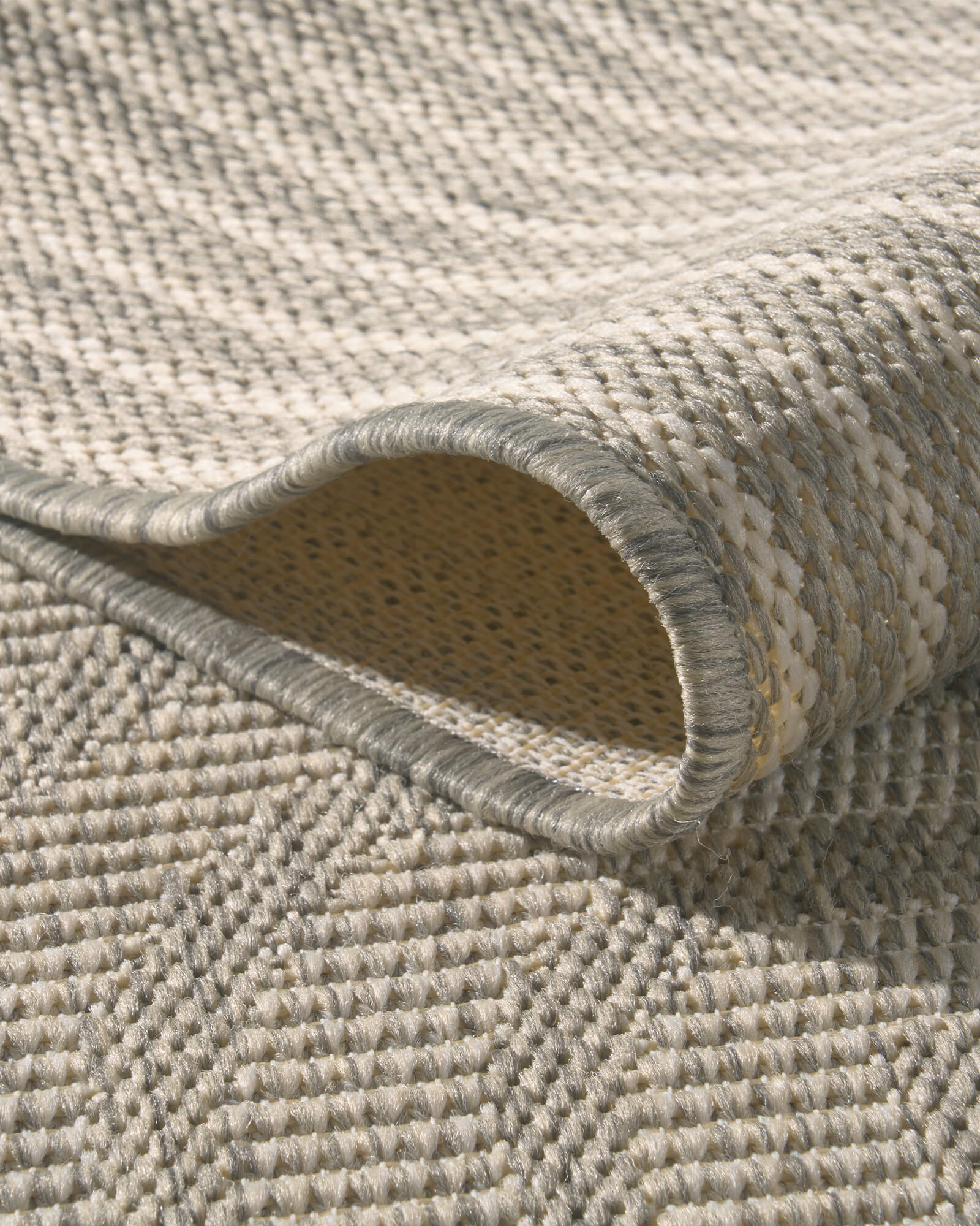 A close-up view of a light gray abstract outdoor rug showing its curved fold, woven back side, raised geometric line texture, and bound edge for durable patio, deck, and porch use.