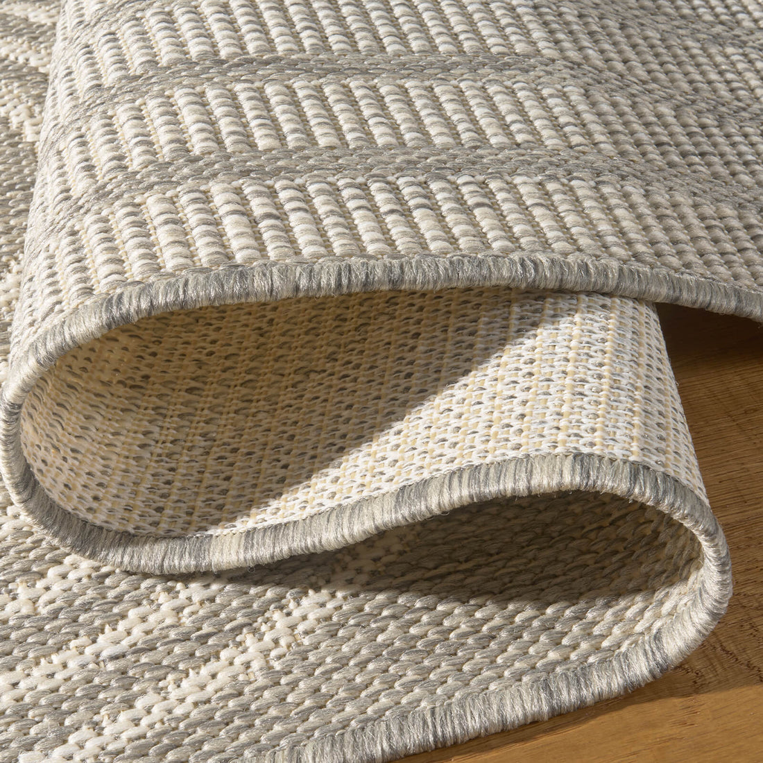 A close-up view of a light gray abstract outdoor rug showing its folded corner, woven backing, bound edge, and raised arch stripe texture for durable patio, deck, and porch use.