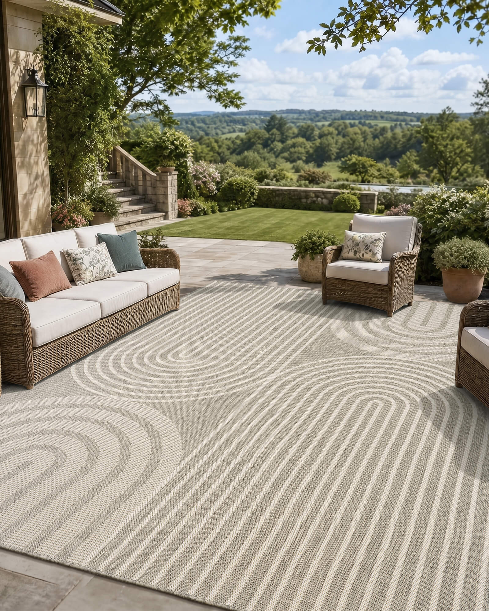A light gray abstract outdoor rug with a modern geometric arch line pattern adds soft texture to a scenic patio lounge with wicker seating, garden greenery, open views, and weather-resistant outdoor style.