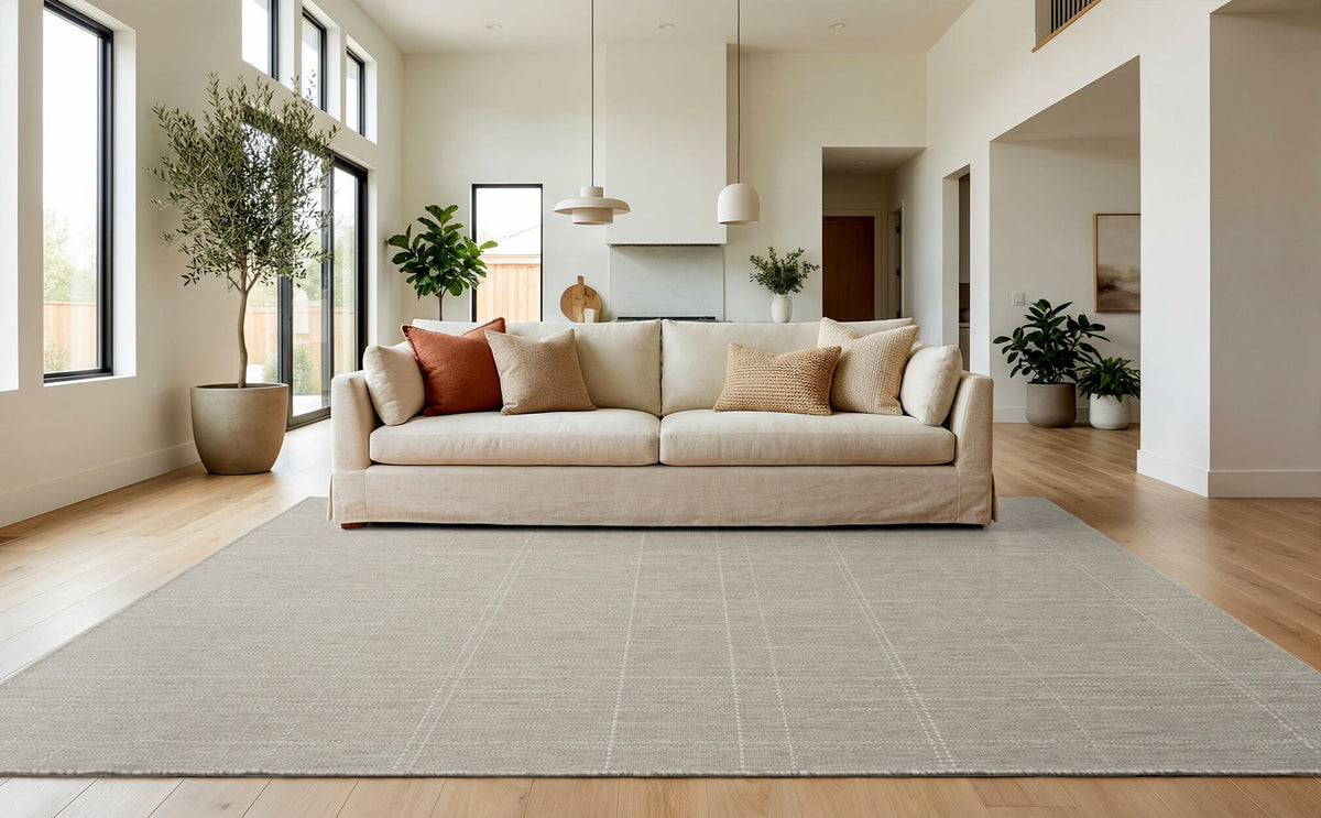 A light gray grid indoor outdoor area rug adds subtle woven texture to an open-plan living room with a cream sofa, tall windows, wood floors, and fresh neutral home decor.