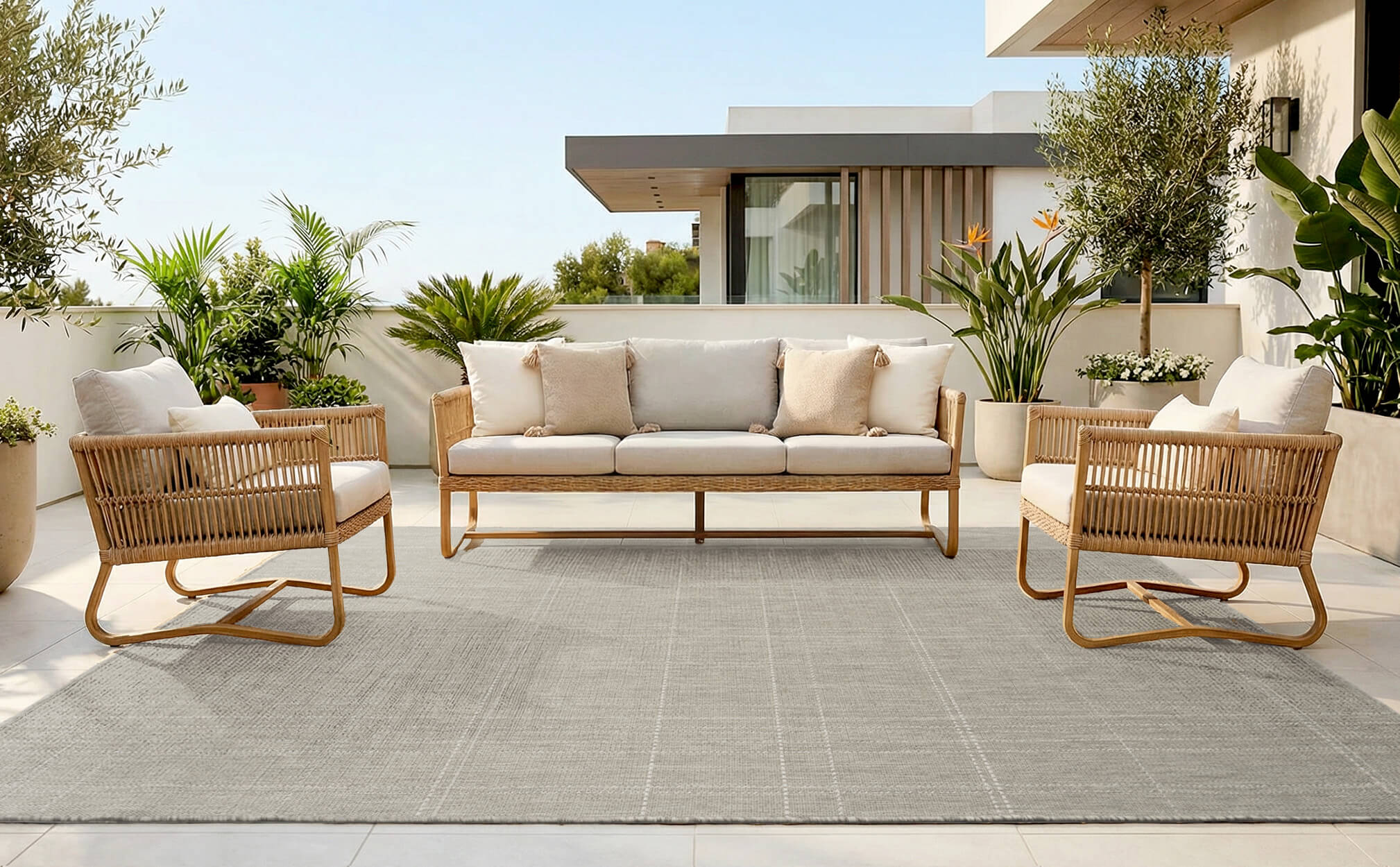 A light gray grid texture outdoor area rug creates a clean terrace seating area with woven chairs, a neutral sofa, tropical greenery, and weather-resistant style for modern patio living.