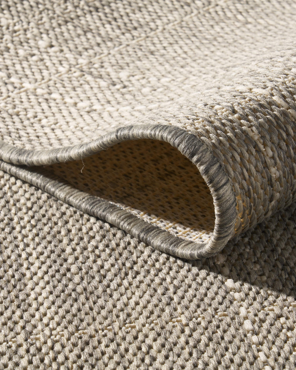 A close-up view of a light gray plain weave outdoor rug showing its folded loop corner, basketweave texture, reinforced edge, and durable woven construction for patios, decks, porches, and outdoor living spaces.