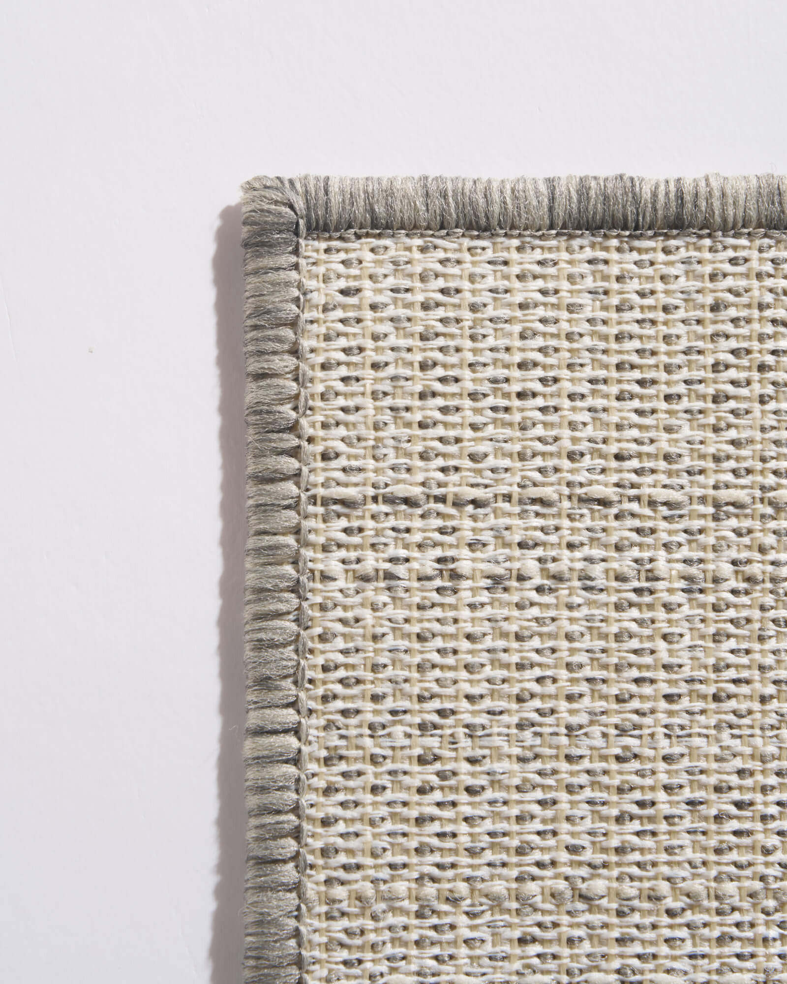 A close-up reverse corner view of a light gray outdoor rug shows its open weave backing, reinforced bound edge, and durable construction for patios, decks, porches, and outdoor entryways.