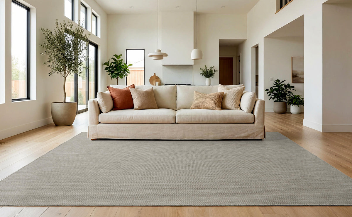 A light gray plain weave indoor outdoor area rug adds subtle woven texture to a bright modern living room with a cream sofa, tall windows, wood floors, and fresh neutral decor.