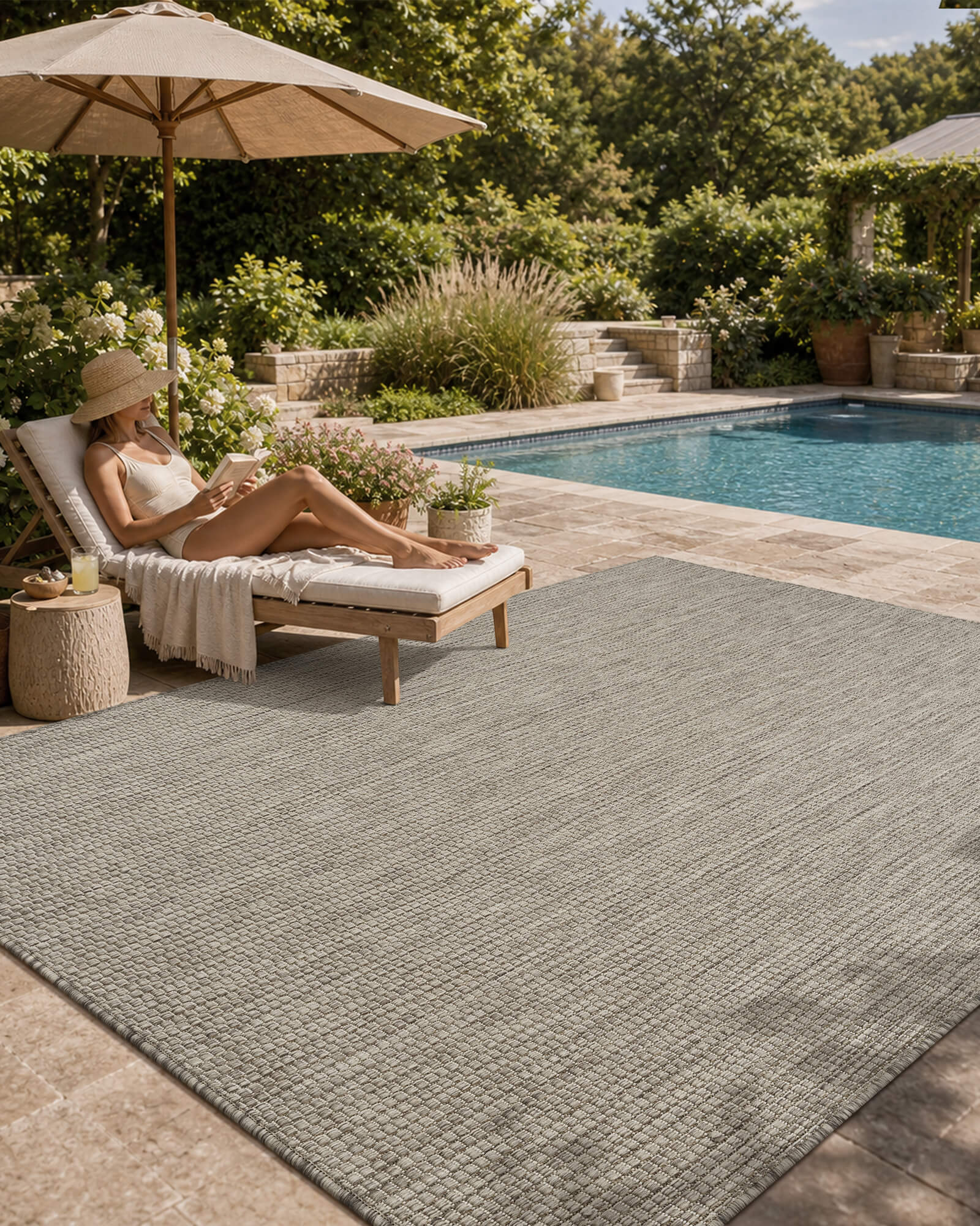 A light gray plain weave outdoor area rug adds clean minimal texture to a poolside patio with a chaise lounge, umbrella shade, garden greenery, and relaxed weather-resistant summer decor.