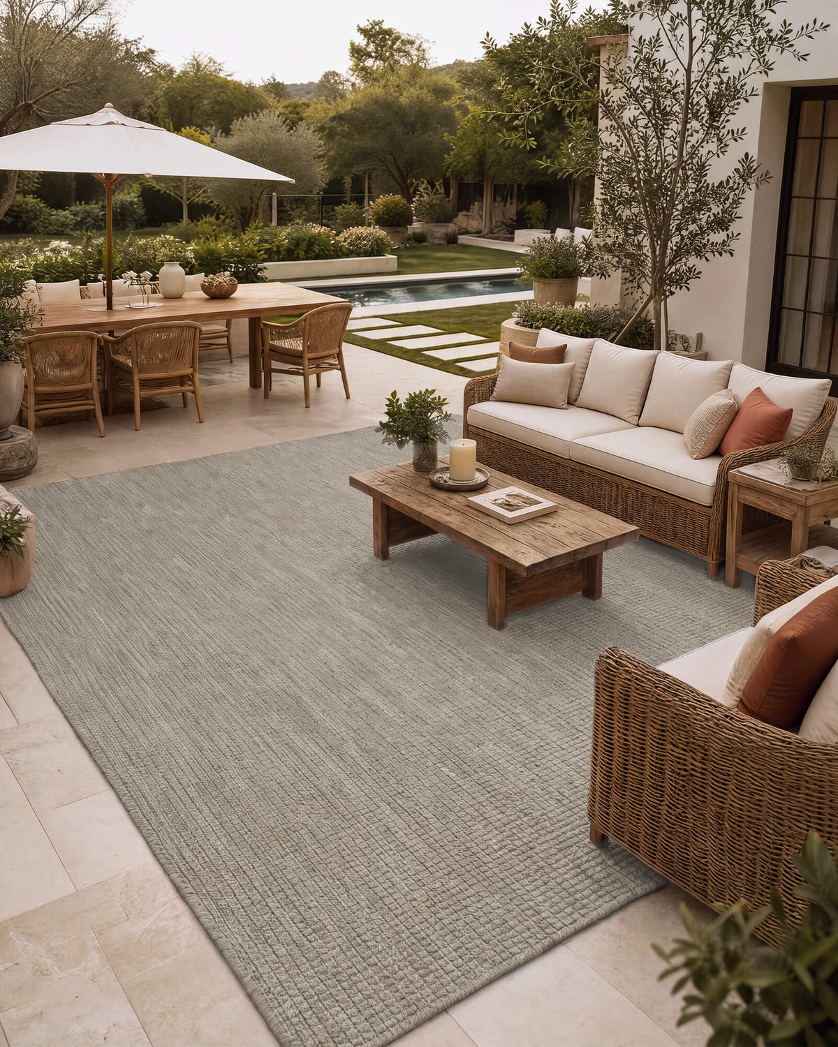 A light gray plain weave outdoor area rug adds clean woven texture to a spacious patio seating area with wicker furniture, a wood coffee table, poolside dining, and relaxed weather-resistant decor.