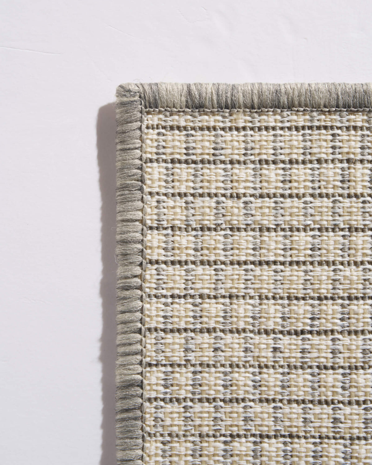 A close-up back corner view of a light gray plain weave outdoor rug shows its open woven backing, finished edge, and durable construction for patios, decks, porches, and outdoor entryways.