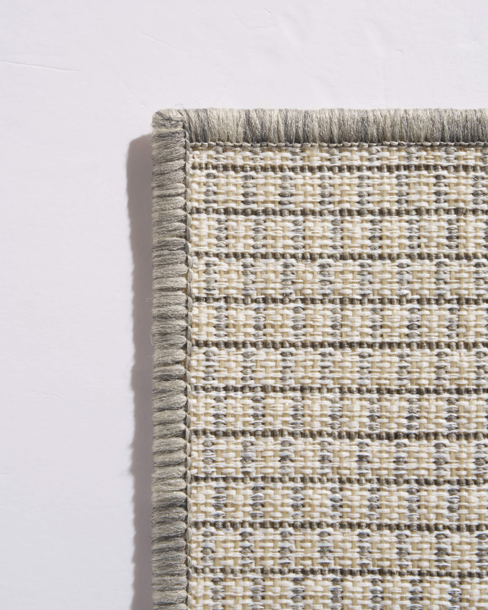 A close-up back corner view of a light gray plain weave outdoor rug shows its open woven backing, finished edge, and durable construction for patios, decks, porches, and outdoor entryways.