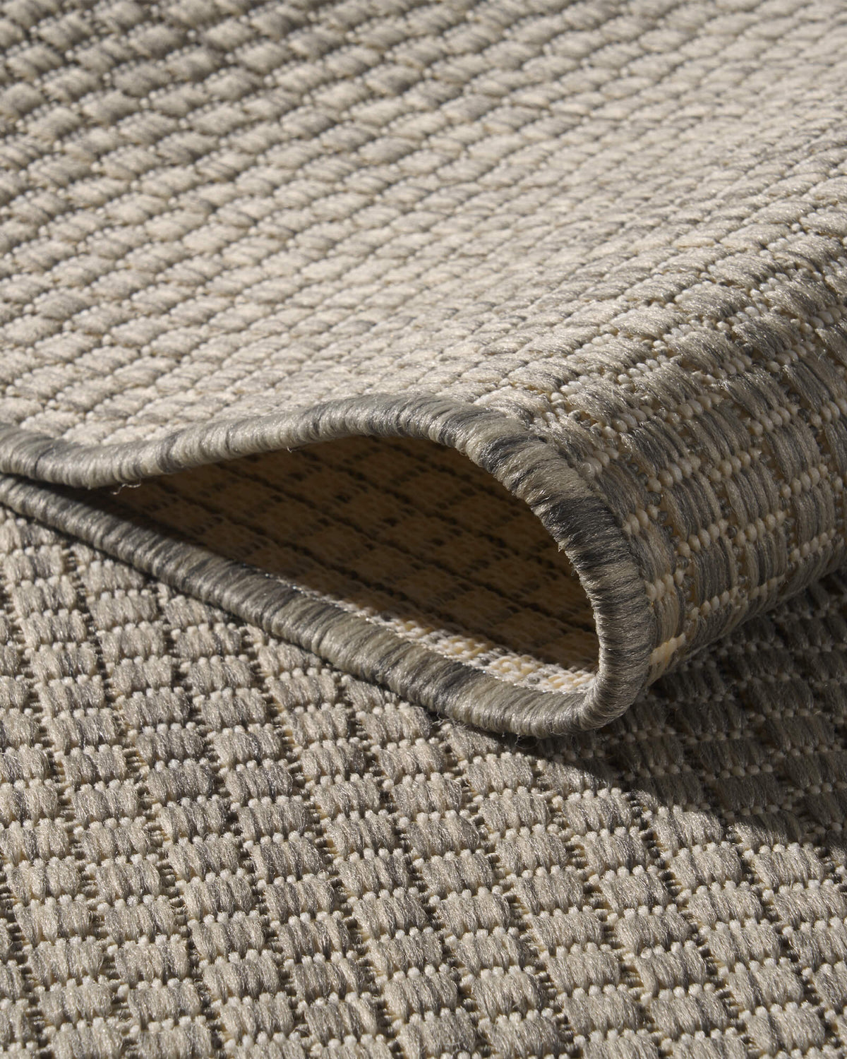 A close-up view of a light gray plain weave outdoor rug showing its looped corner, woven backing, bound edge, and basketweave texture for durable patio, deck, porch, and outdoor living use.