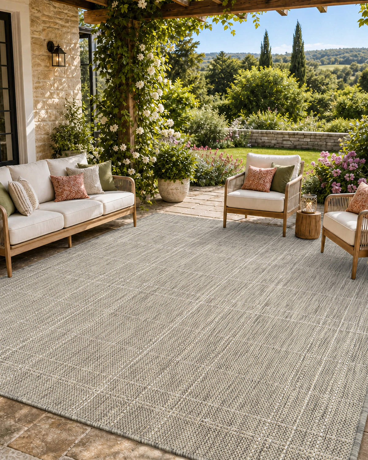 A light gray subtle grid outdoor rug adds clean woven texture to a covered patio lounge with wicker seating, stone flooring, garden greenery, open views, and relaxed weather-resistant style.