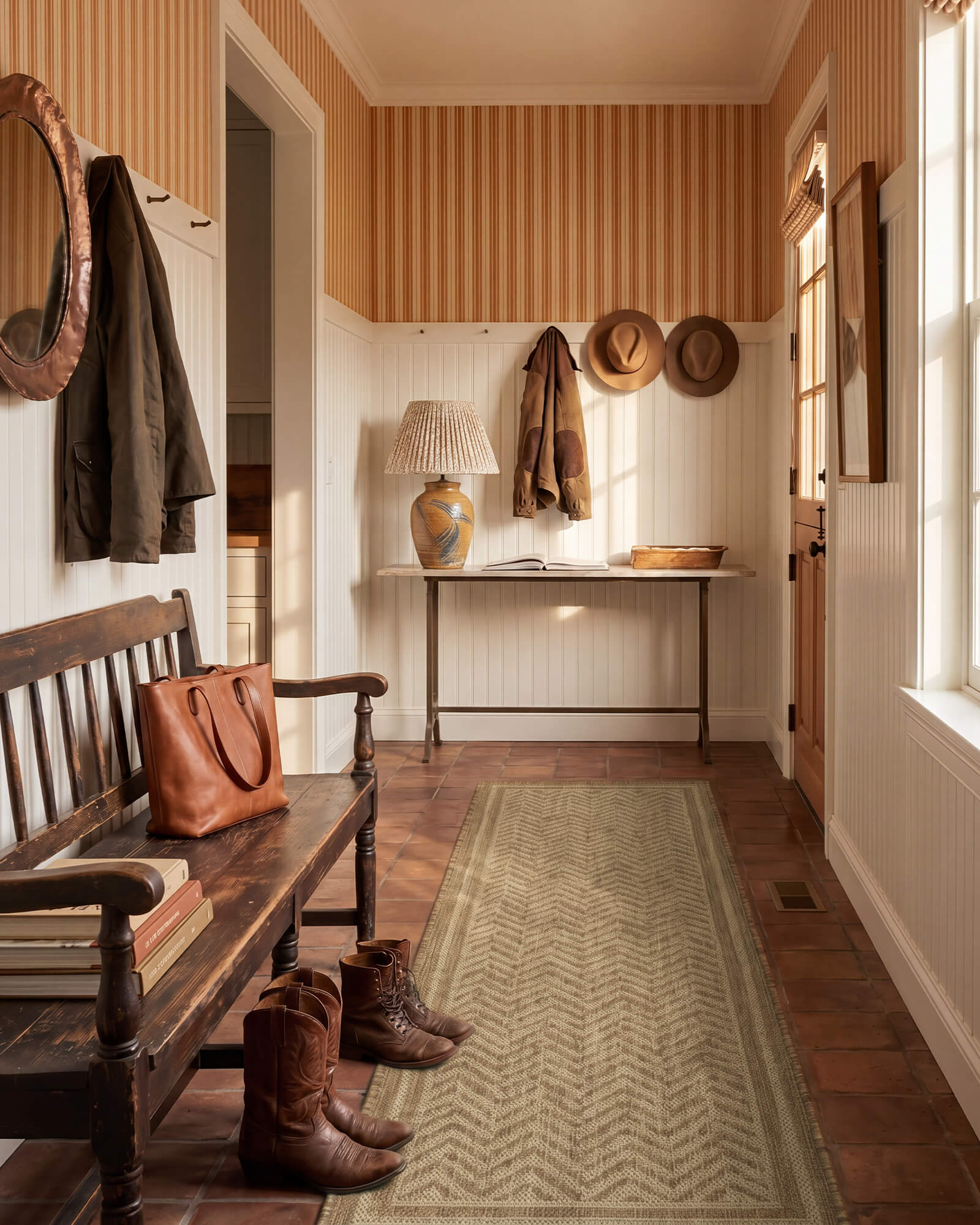 A natural chevron indoor outdoor runner rug adds durable woven texture and easy-care style to an entryway hallway with vintage wood bench, warm sunlight, boots, and classic home decor.